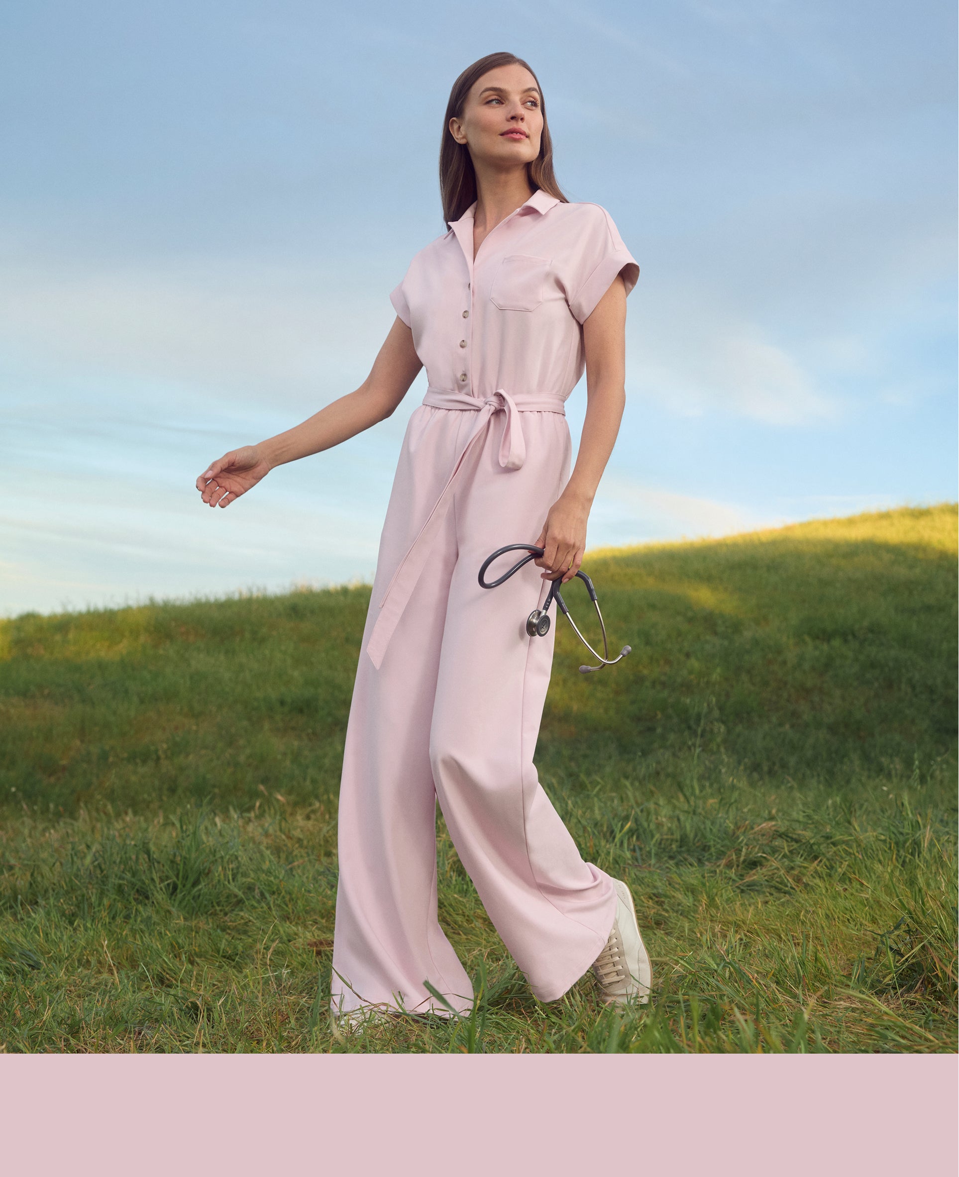 Woman in a rose quartz Jaanuu short-sleeve belted jumpsuit walking across a grassy hill holding a stethoscope, with a rose quartz color band across the bottom