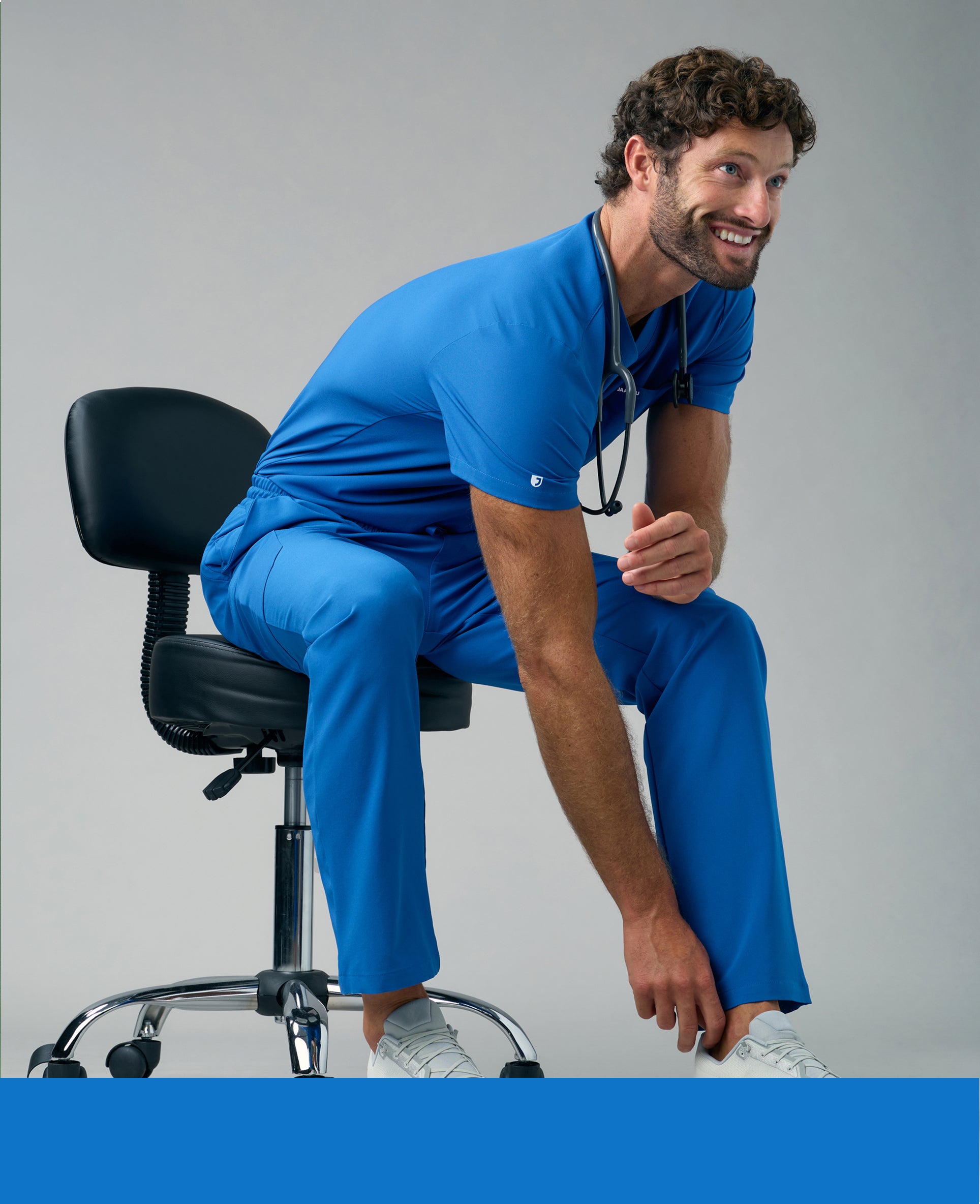 Smiling man in a royal blue Jaanuu V-neck scrub top and scrub pants sitting on a rolling stool with a stethoscope around his neck, reaching for his white sneaker, with a royal blue color band across the bottom