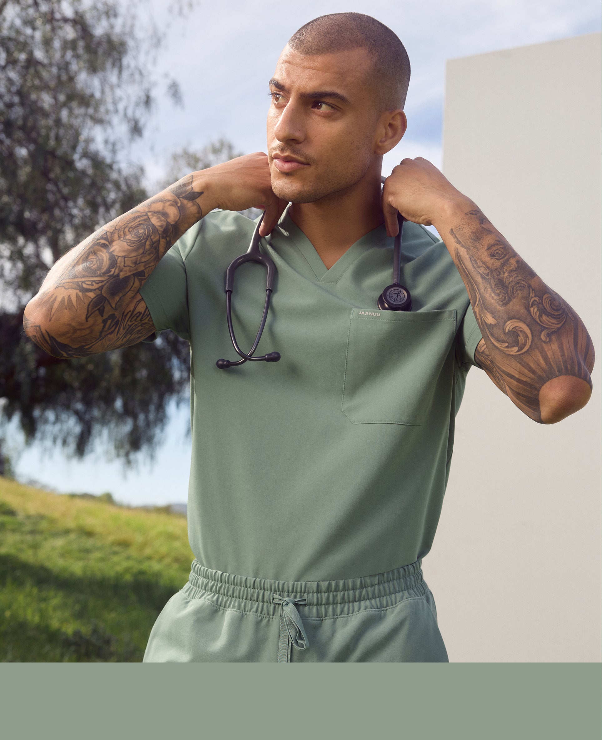 Man in a sage Jaanuu V-neck scrub top and scrub pants adjusting the stethoscope around his neck, with a tree and hillside behind him, with a sage color band across the bottom