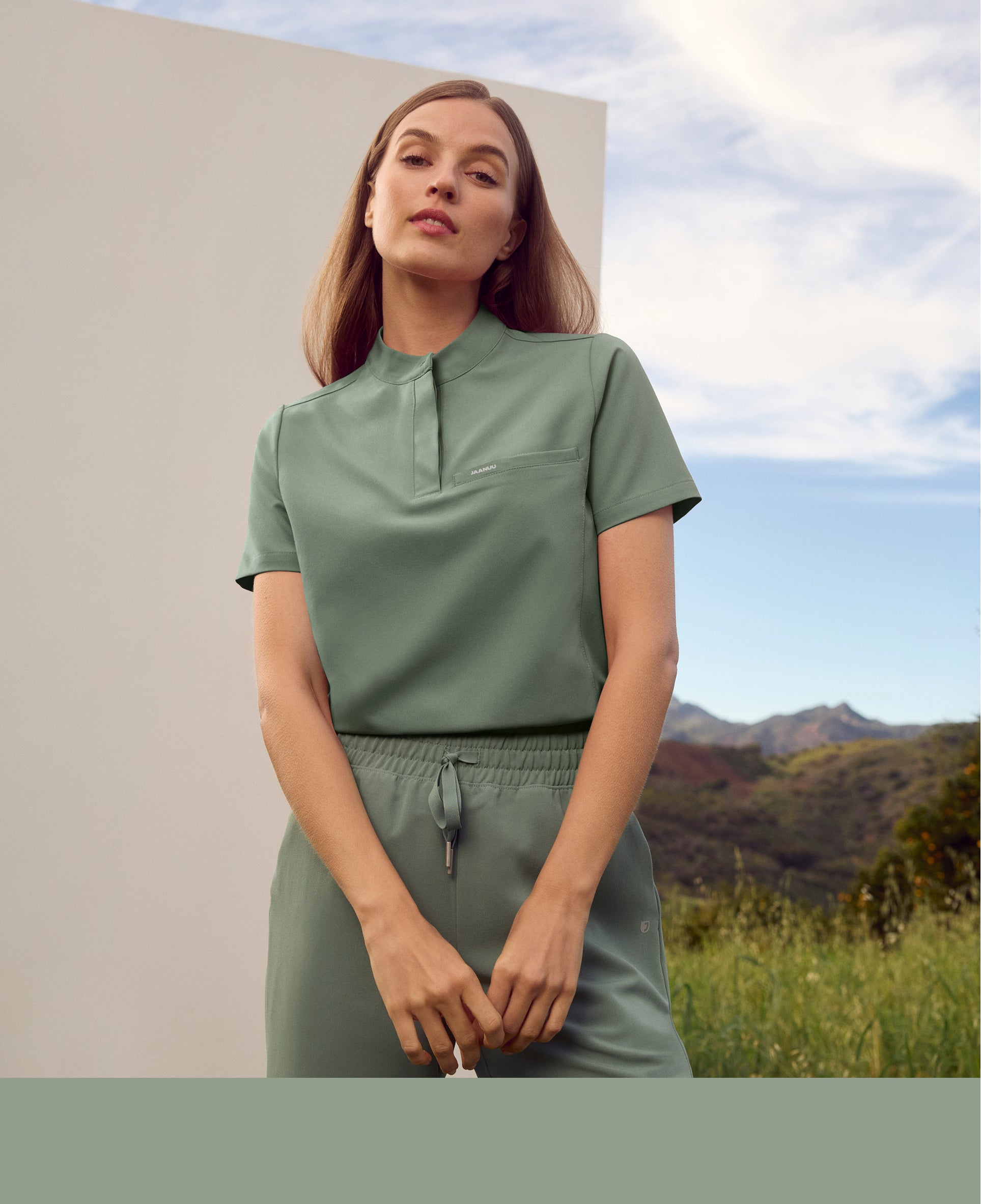 Woman in a sage Jaanuu stand-collar scrub top and jogger scrub pants standing in front of a white wall with green hills and sky behind her, with a sage color band across the bottom
