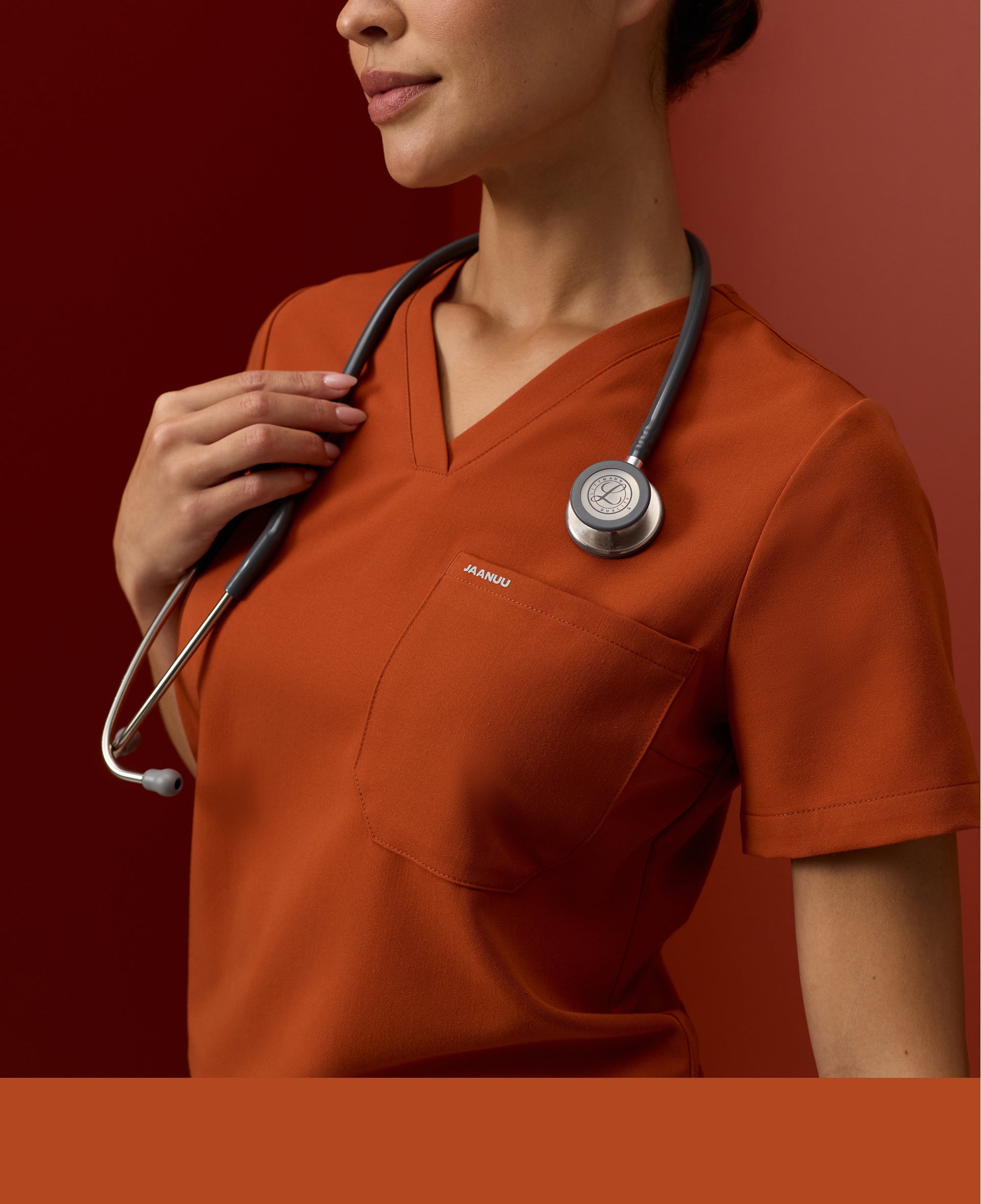 Close-up of a woman in a sienna Jaanuu V-neck scrub top holding the stethoscope around her neck against a dark red backdrop, with a sienna color band across the bottom