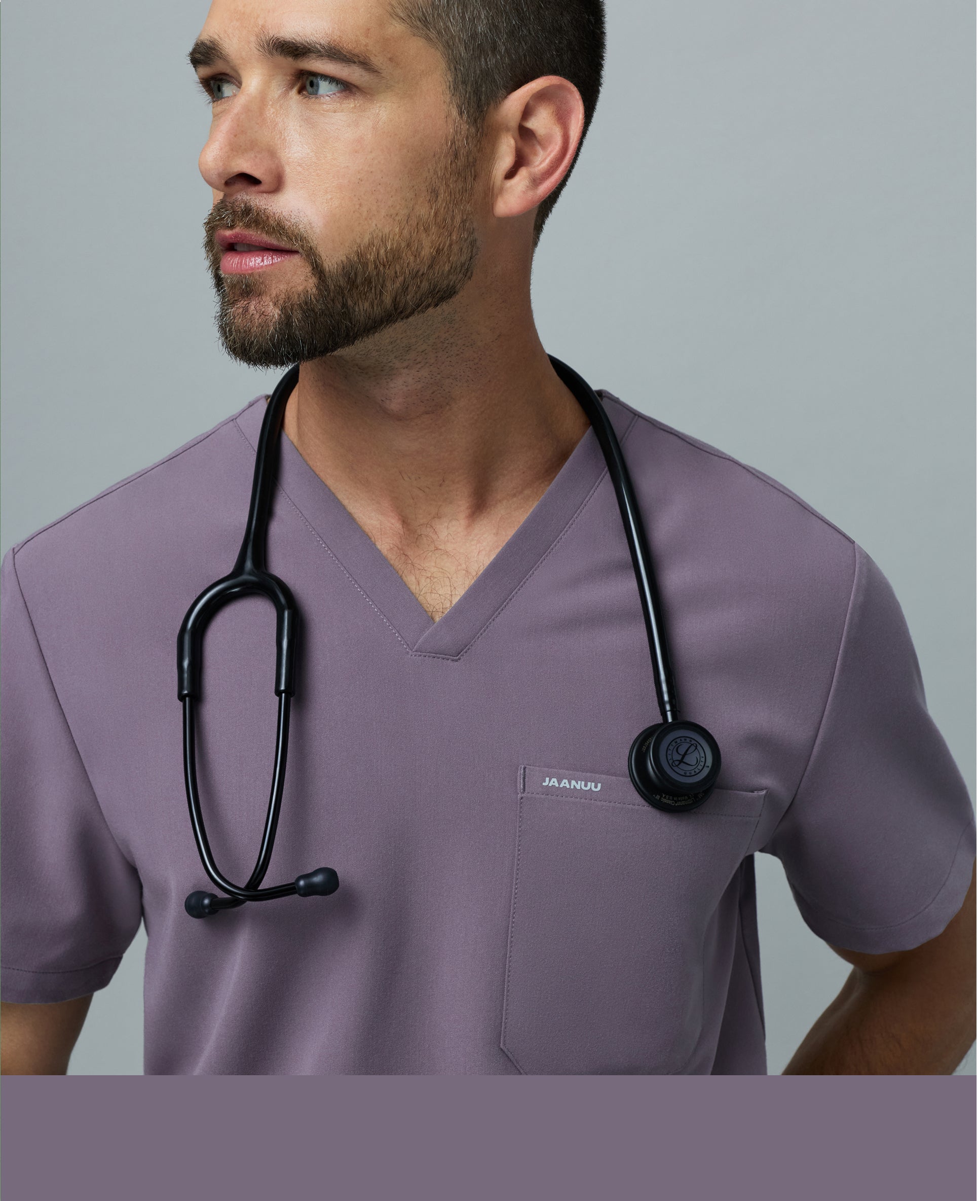Close-up of a man in a titanium Jaanuu V-neck scrub top with a stethoscope around his neck looking to the side against a gray backdrop, with a titanium color band across the bottom