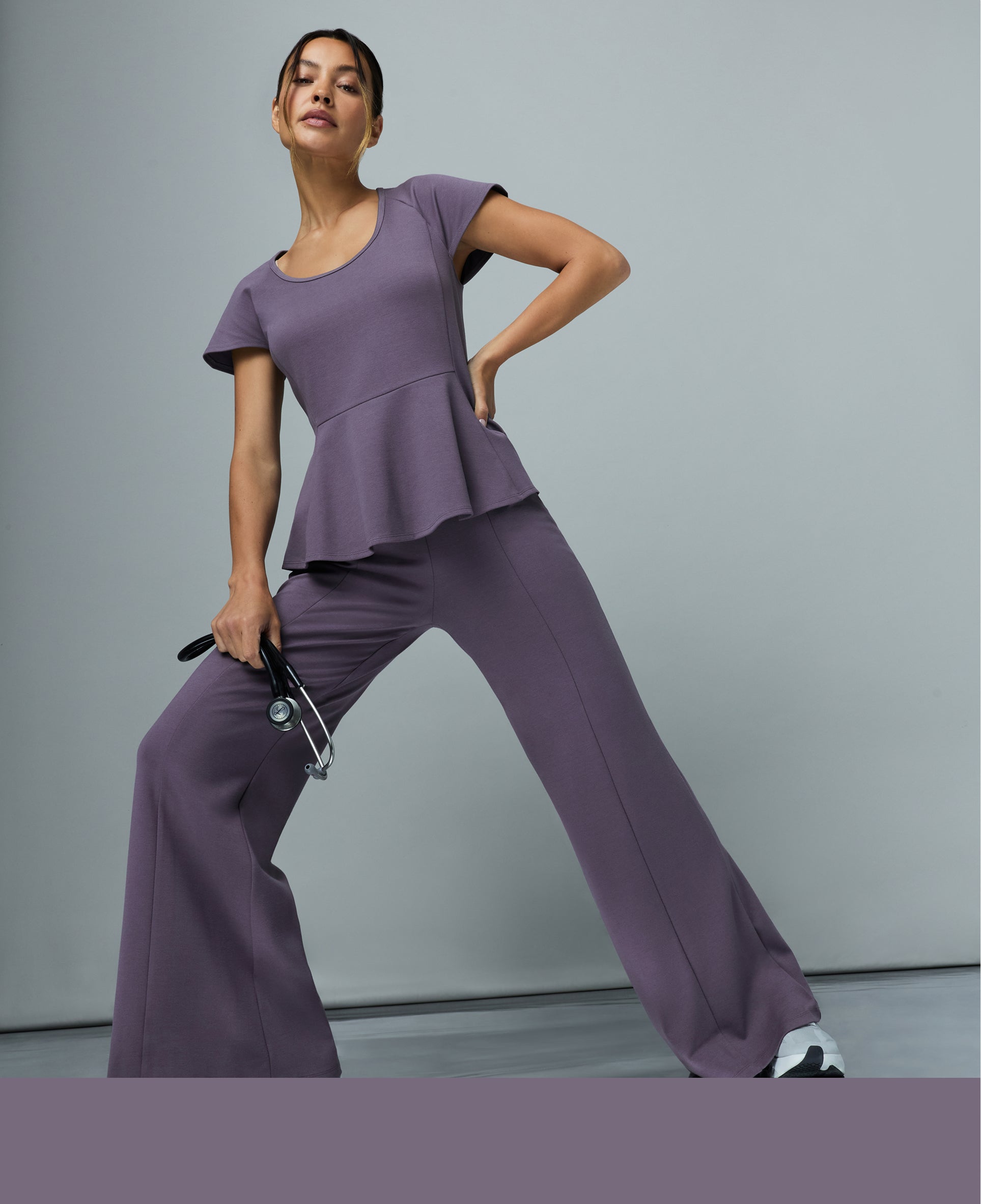 Woman in a titanium Jaanuu scoop-neck peplum scrub top and wide-leg scrub pants, standing with one hand on her hip and a stethoscope in the other against a gray backdrop, with a titanium color band across the bottom