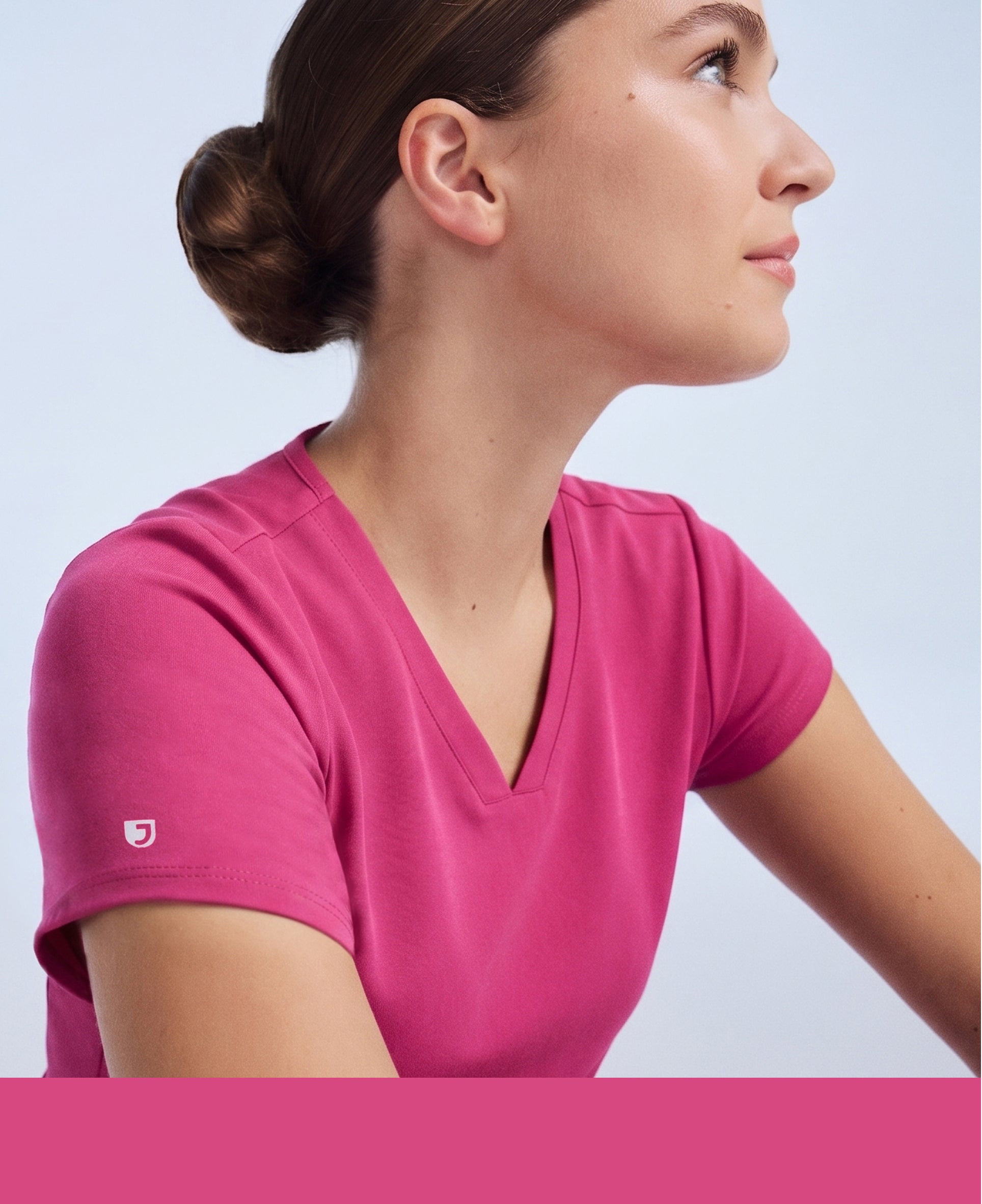 Profile of a woman in a warrior pink Jaanuu V-neck scrub top looking up against a pale blue backdrop, with a warrior pink color band across the bottom