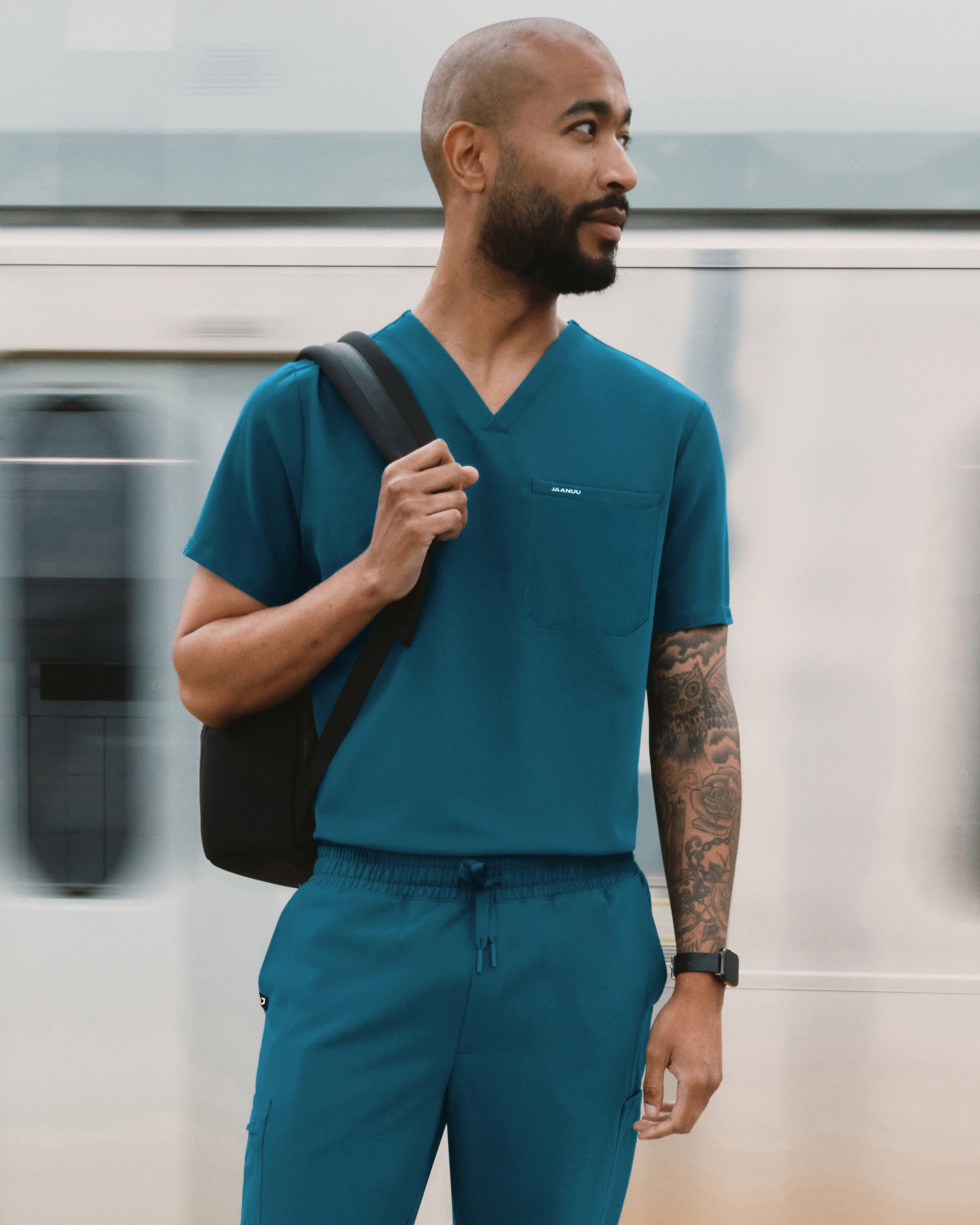 Man in a teal Jaanuu V-neck scrub top and jogger scrub pants with a backpack over one shoulder, standing in front of a passing train