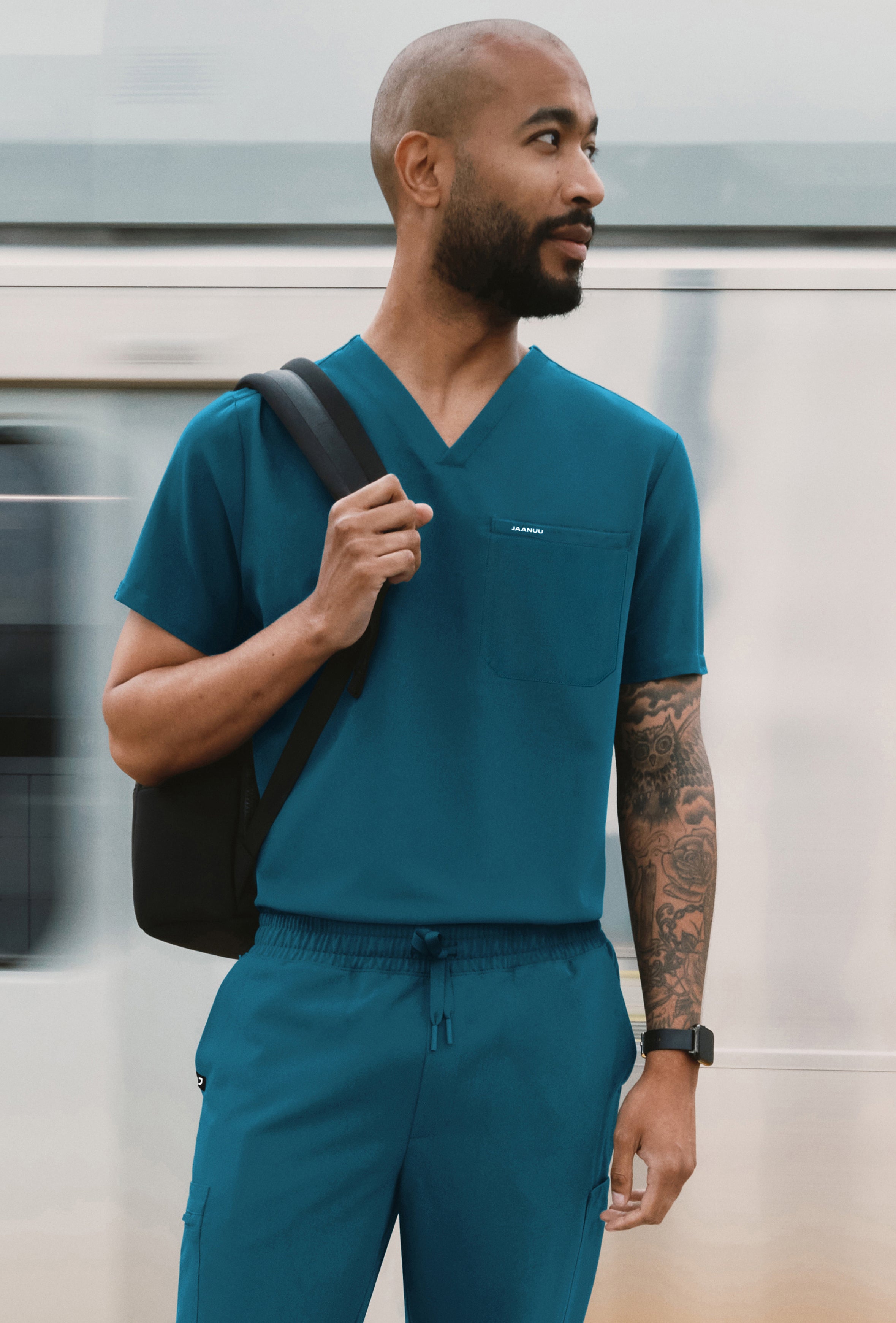 Man in a teal Jaanuu V-neck scrub top and jogger scrub pants with a backpack over one shoulder, standing in front of a passing train