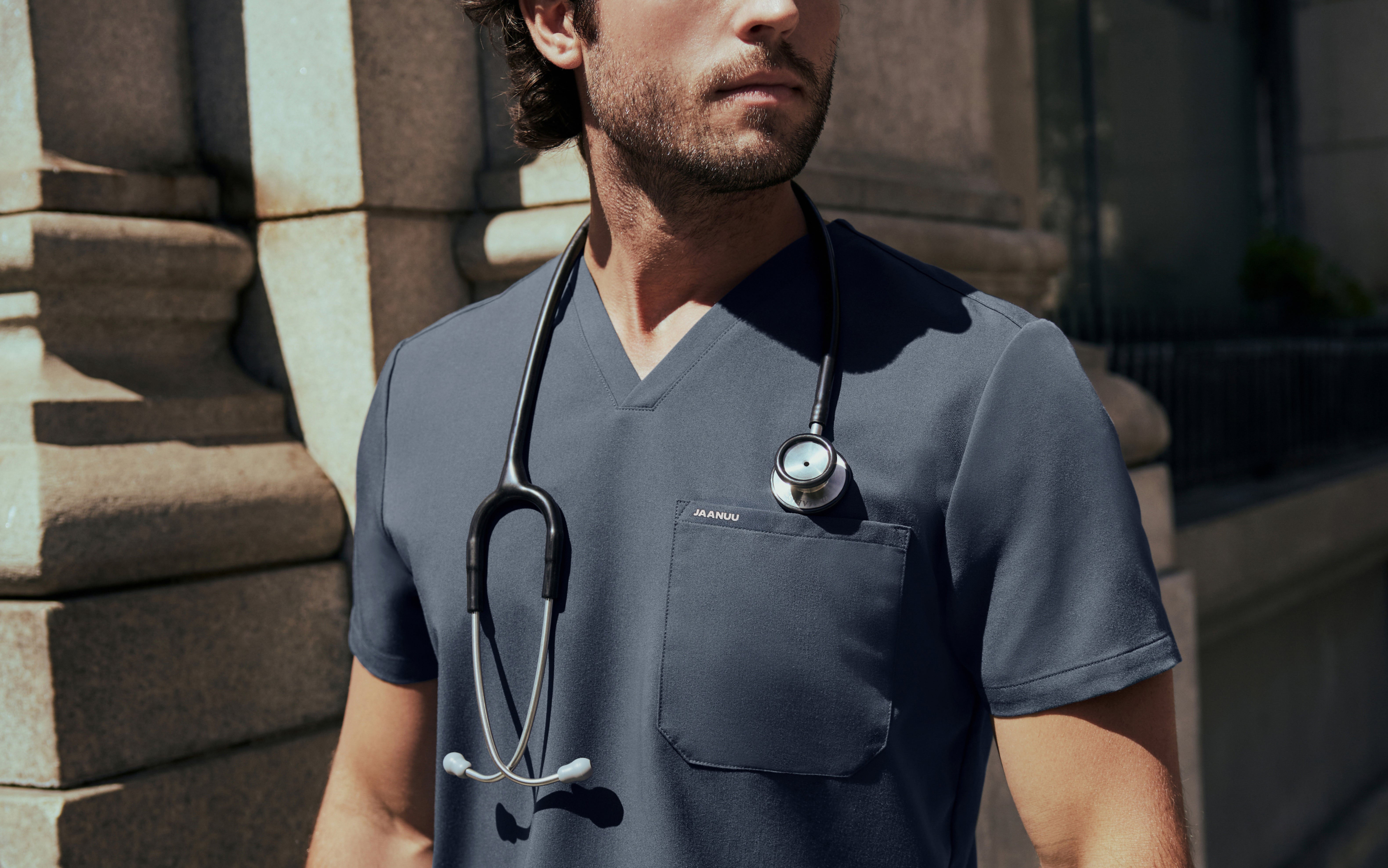 Close-up of a man in a charcoal gray Jaanuu V-neck scrub top with a stethoscope around his neck standing beside a stone building in sunlight
