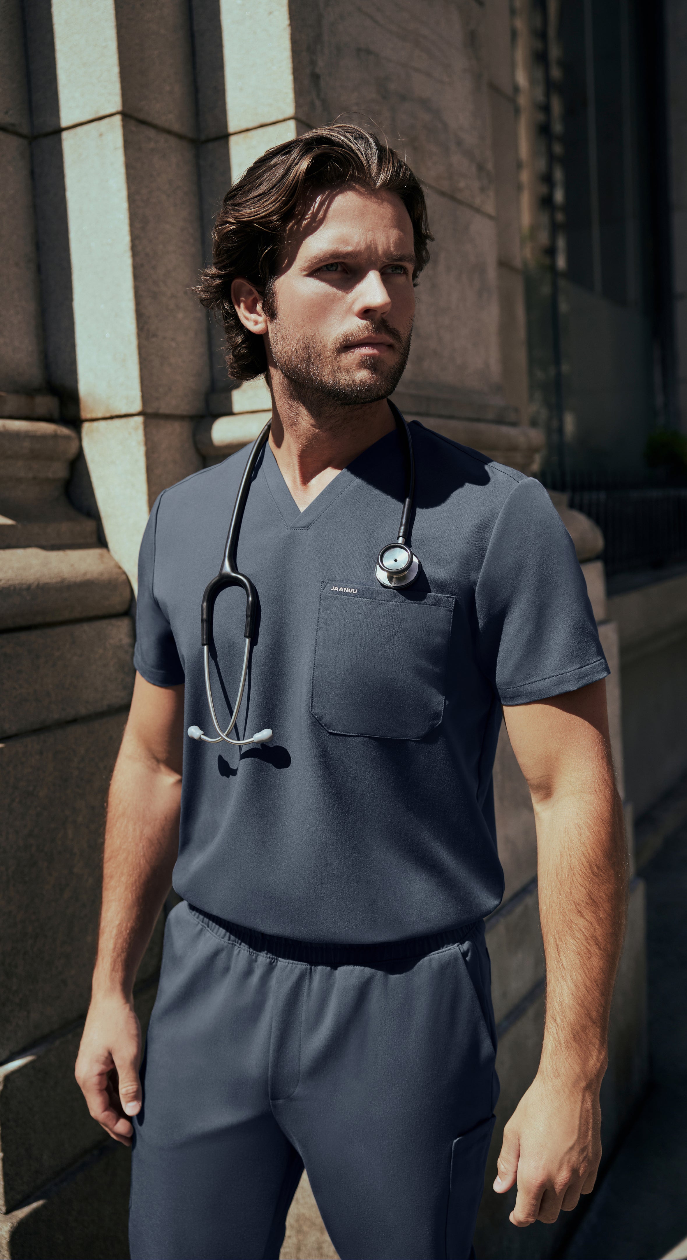 Man in a charcoal gray Jaanuu V-neck scrub top and scrub pants with a stethoscope around his neck standing beside a stone building in sunlight