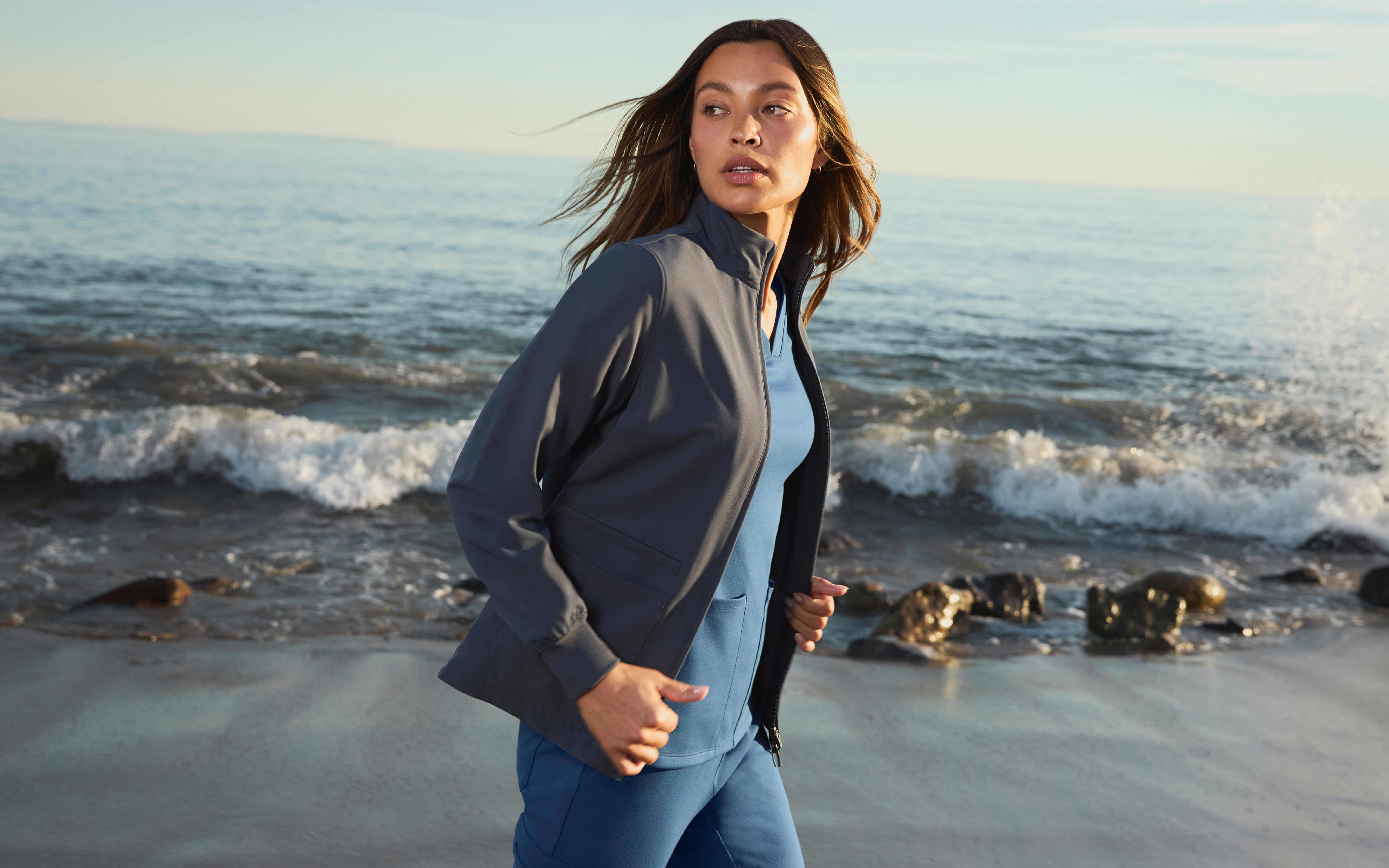 Woman in a charcoal gray Jaanuu zip-up scrub jacket over a blue scrub top and cargo scrub pants walking along a beach with waves behind her