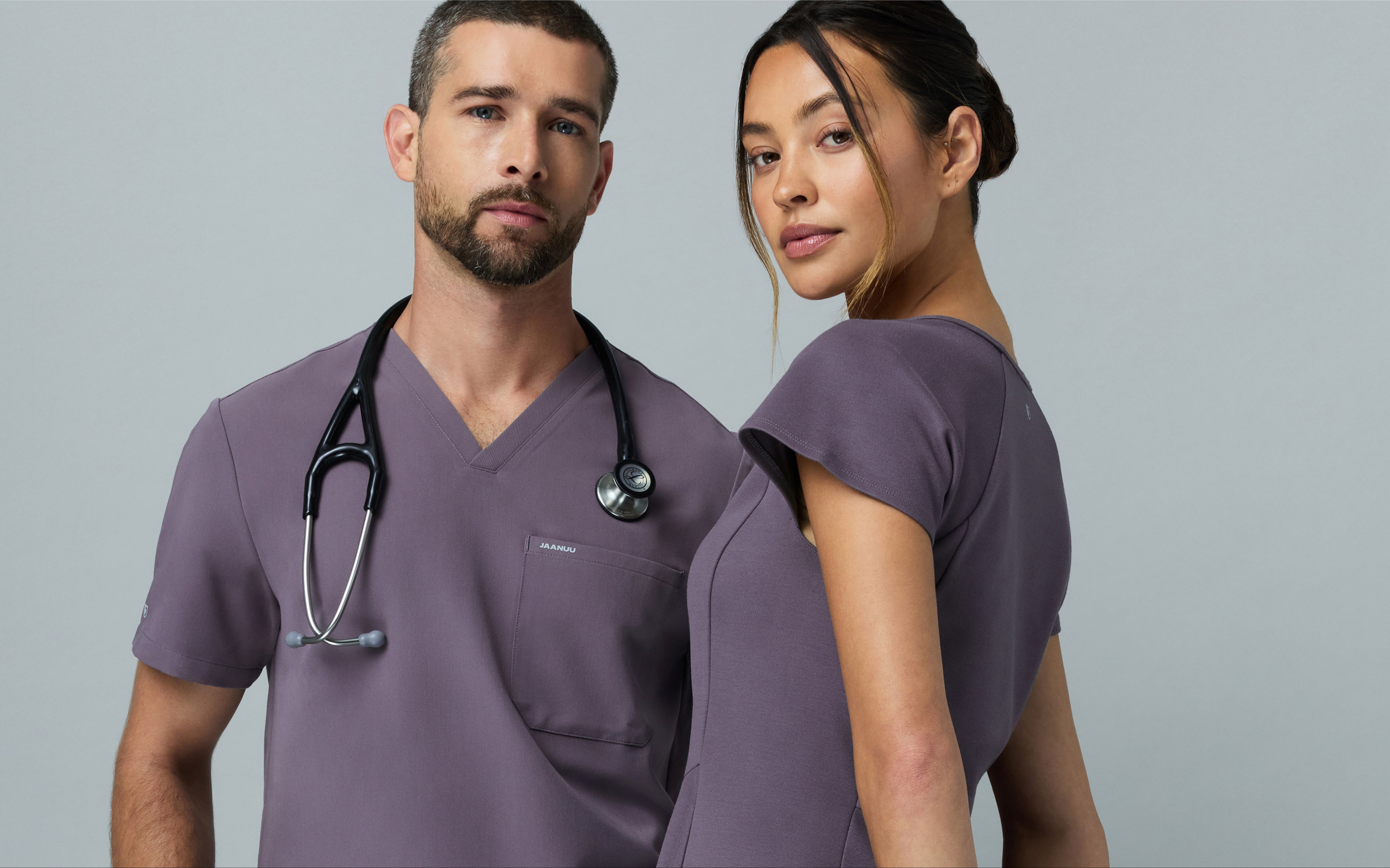 Man and woman in matching muted purple Jaanuu scrub sets standing back to back against a gray backdrop, he with a stethoscope around his neck