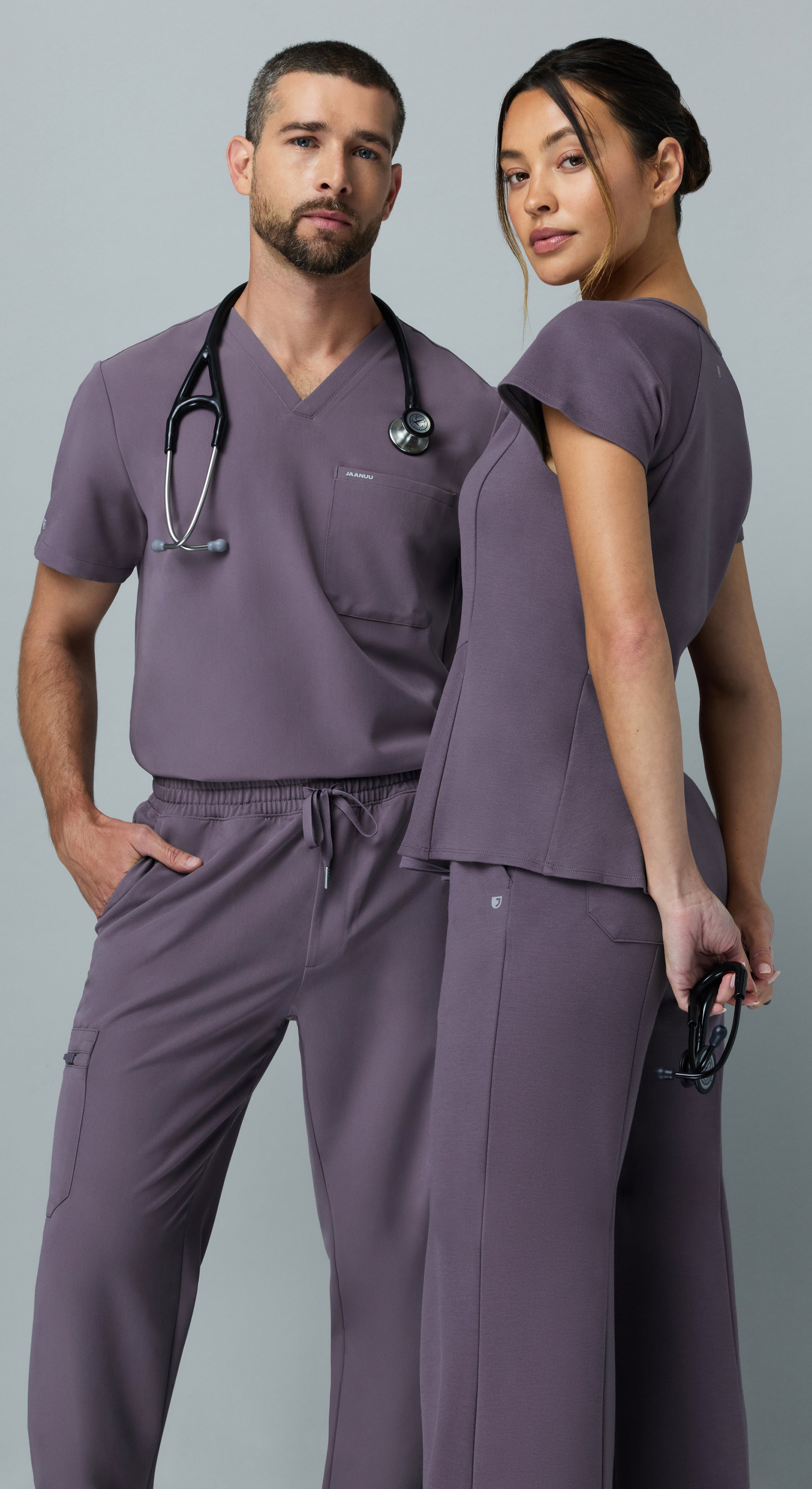 Man and woman in matching muted purple Jaanuu scrub sets standing back to back against a gray backdrop, he with a stethoscope around his neck and she holding one at her side