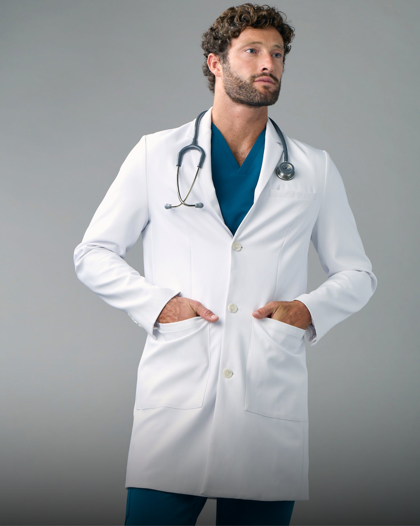 Man in a white lab coat over scrubs with a stethoscope around his neck and his hands in the coat pockets