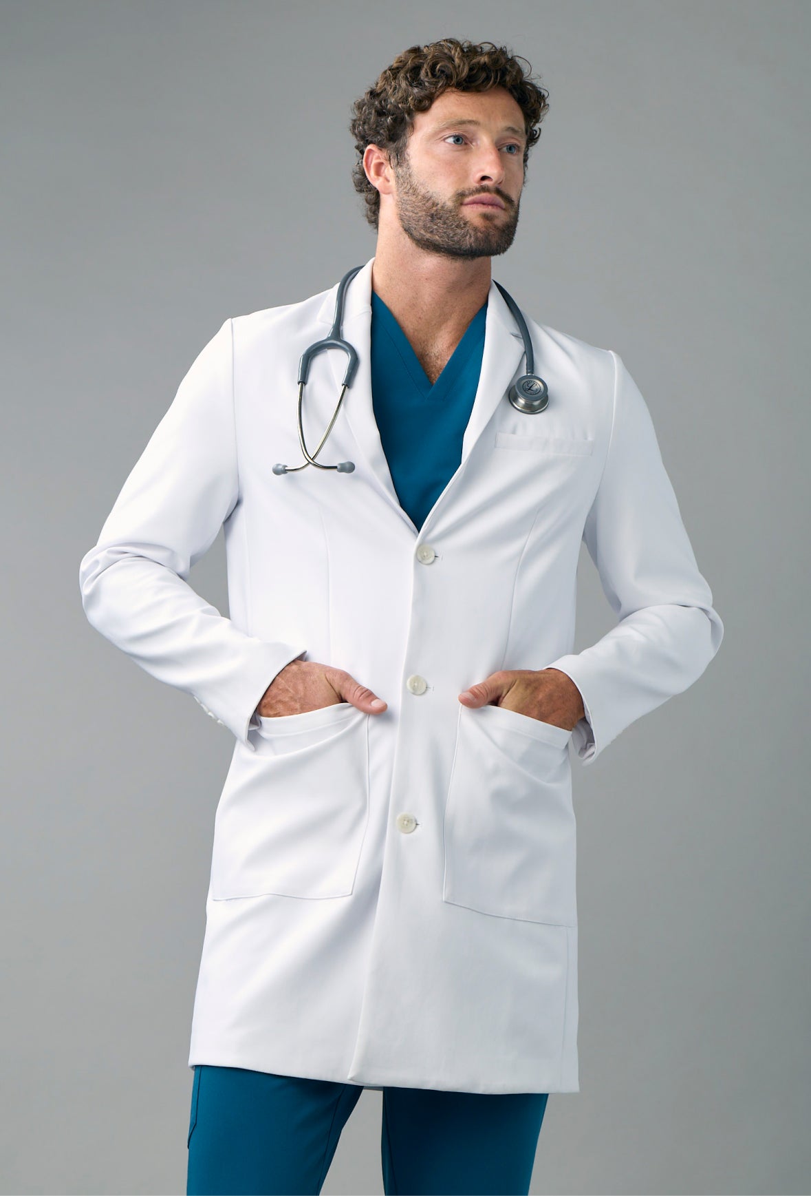 Man in a white lab coat over scrubs with a stethoscope around his neck and his hands in the coat pockets