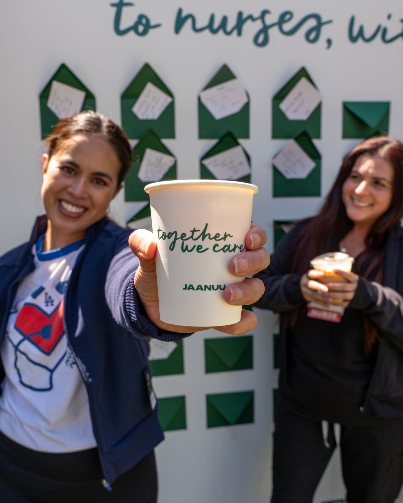 Two women holding JAANUU Together We Care cups in front of a wall of green envelopes with notes to nurses