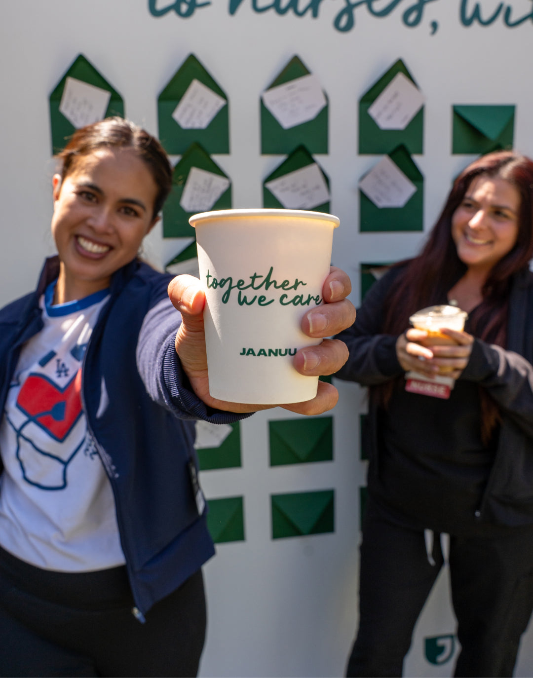 Two women holding JAANUU Together We Care cups in front of a wall of green envelopes with notes to nurses