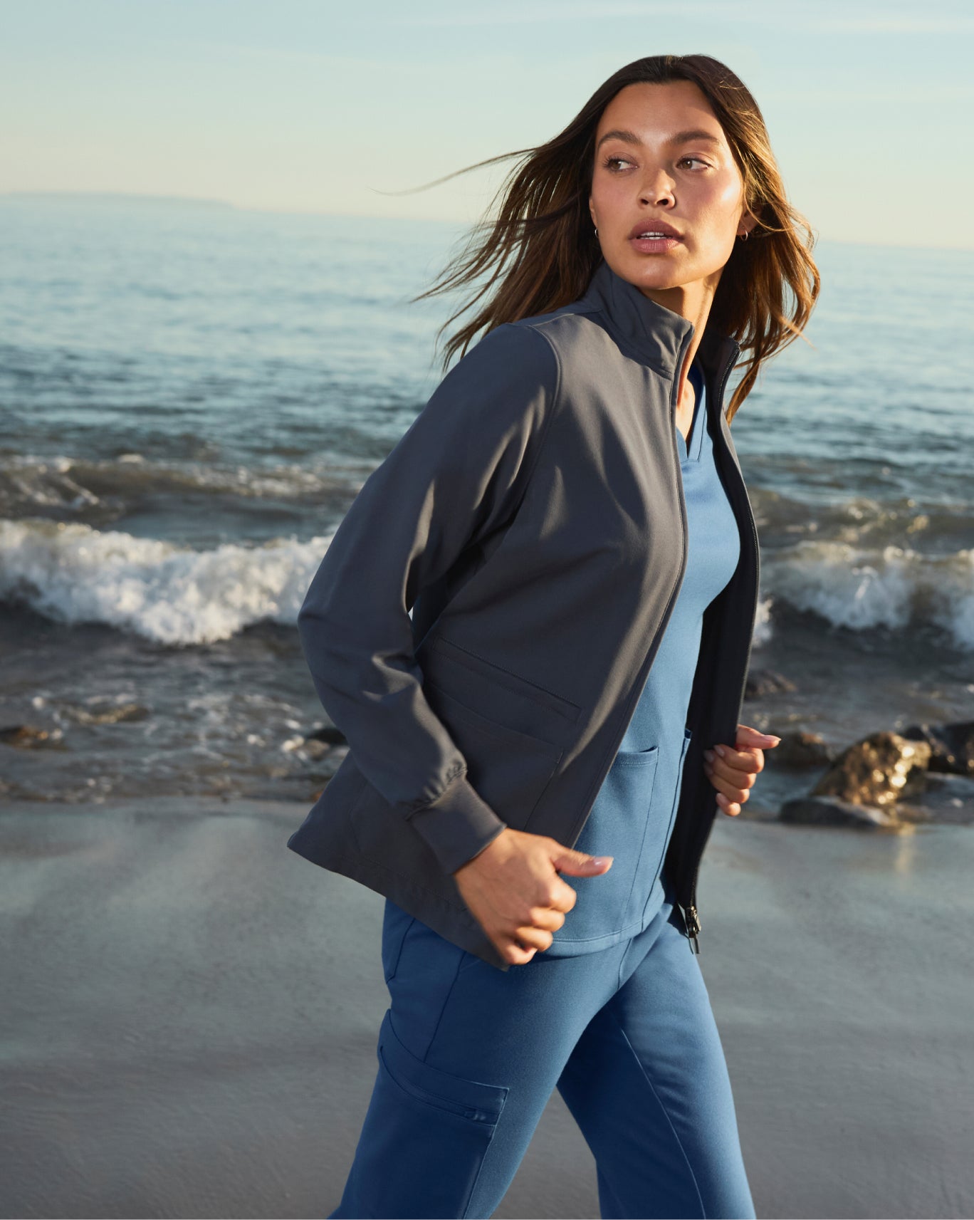 Woman in a dark zip jacket on a beach with waves breaking behind her