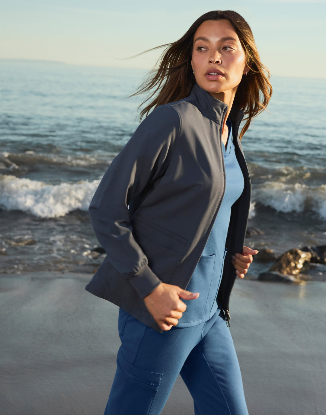 Woman in a dark zip jacket on a beach with waves breaking behind her