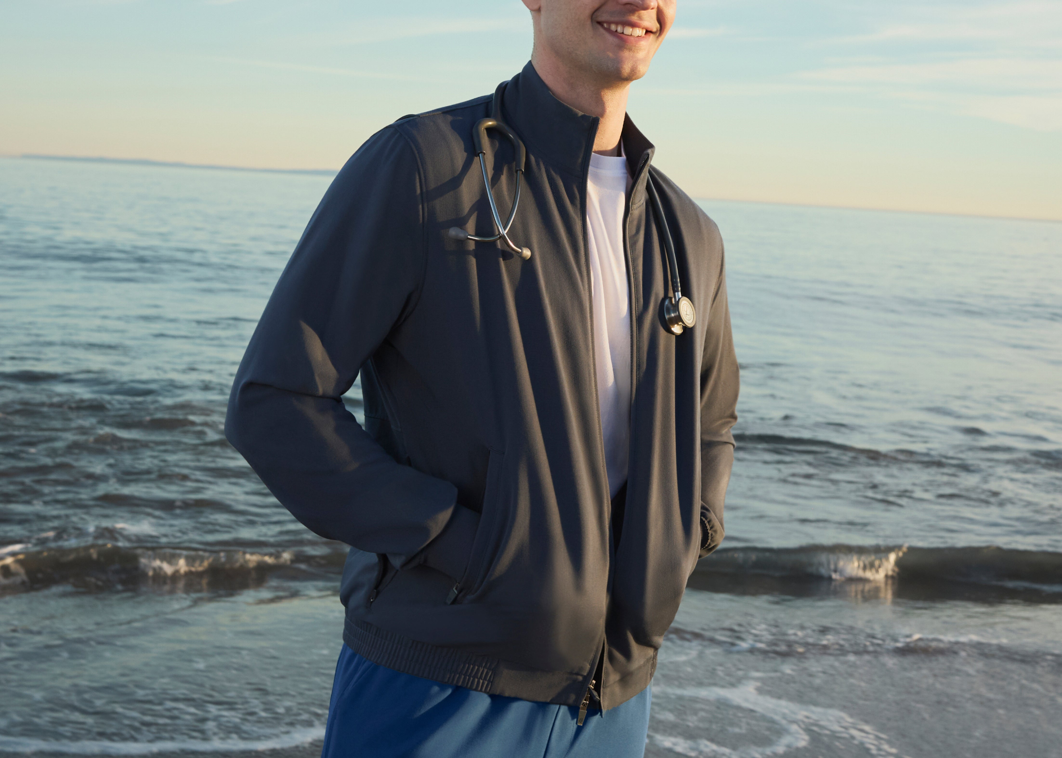 Smiling man in a charcoal zip-up scrub jacket over a white T-shirt, a stethoscope around his neck and his hands in the jacket pockets, on a beach at sunset