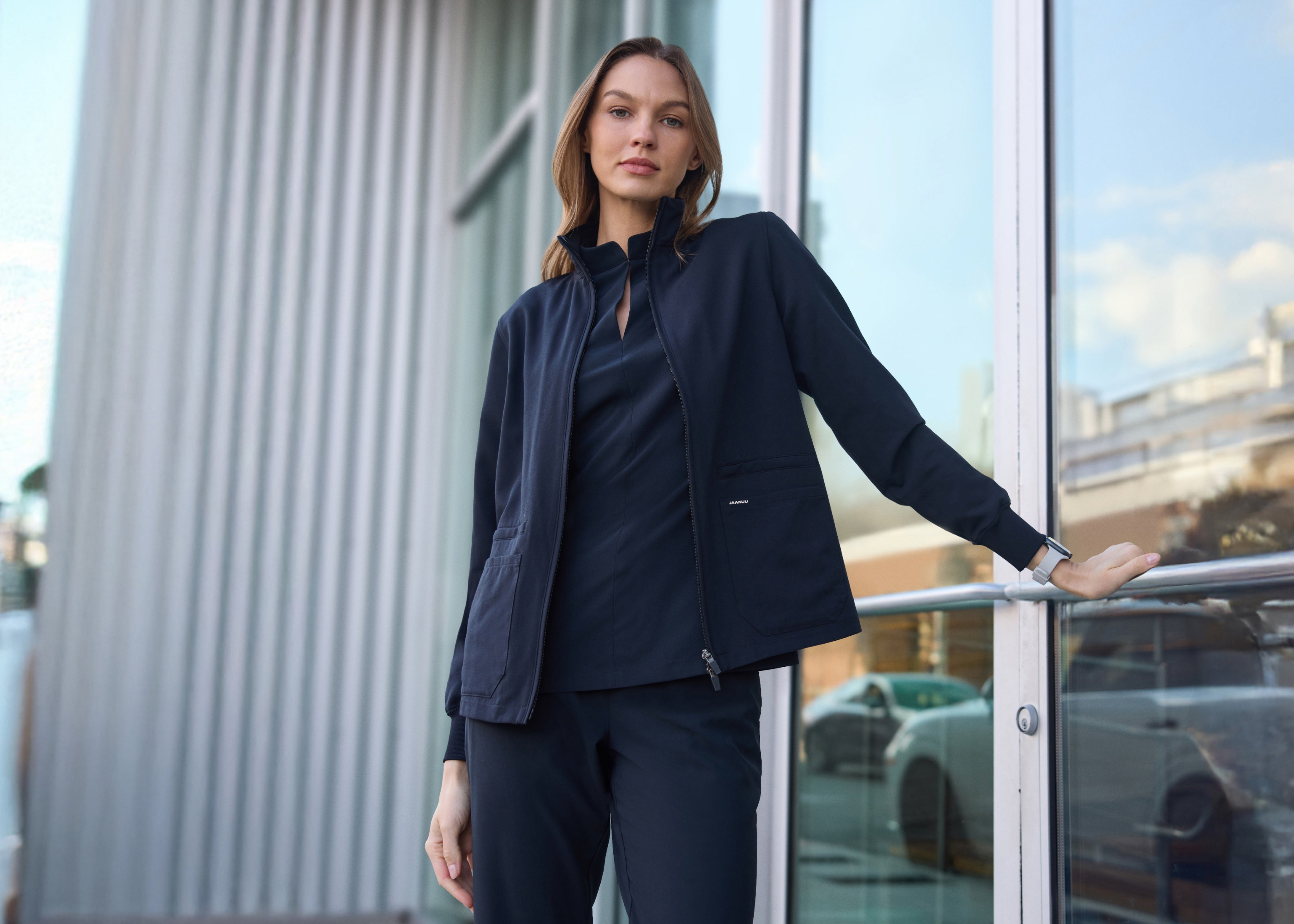 Woman in a navy zip-up scrub jacket over a navy scrub top and scrub pants, one hand on the rail of a glass door outside a building
