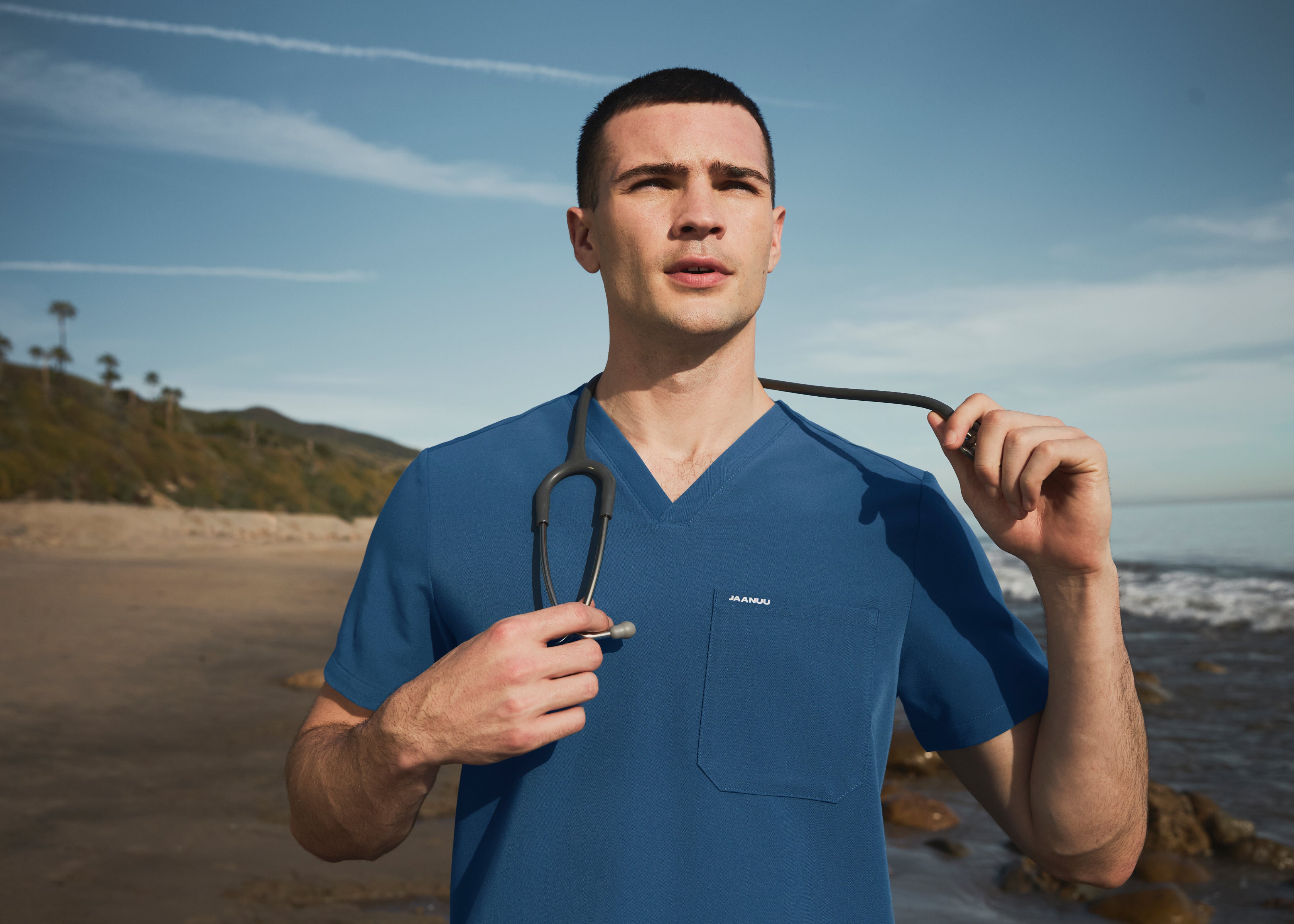 Man in a blue V-neck scrub top with JAANUU on the chest pocket, holding a stethoscope around his neck on a beach
