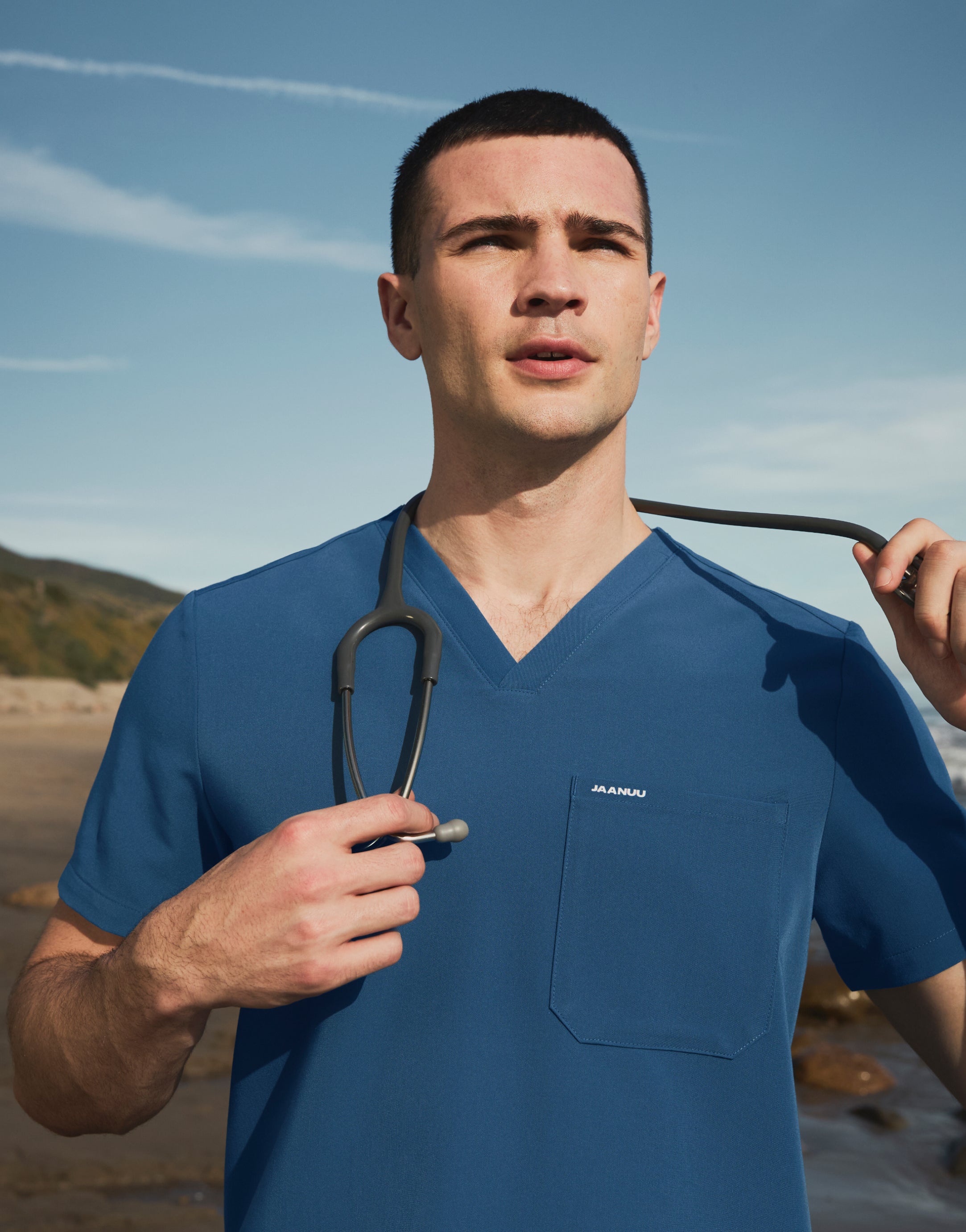 Man in a blue V-neck scrub top with JAANUU on the chest pocket, holding a stethoscope around his neck on a beach under a blue sky