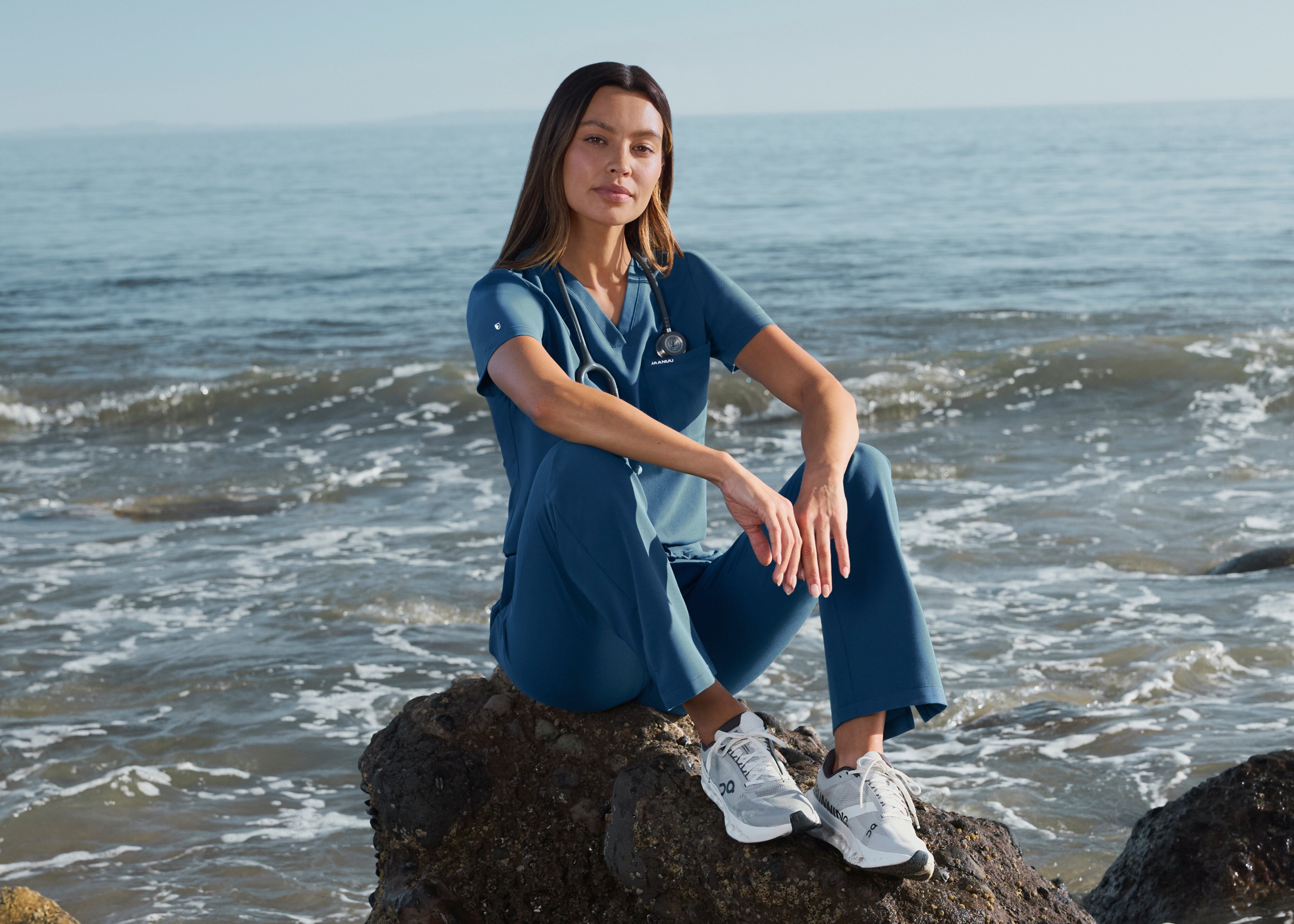 Woman in a blue scrub top and scrub pants with a stethoscope around her neck, sitting on a rock at the edge of the ocean