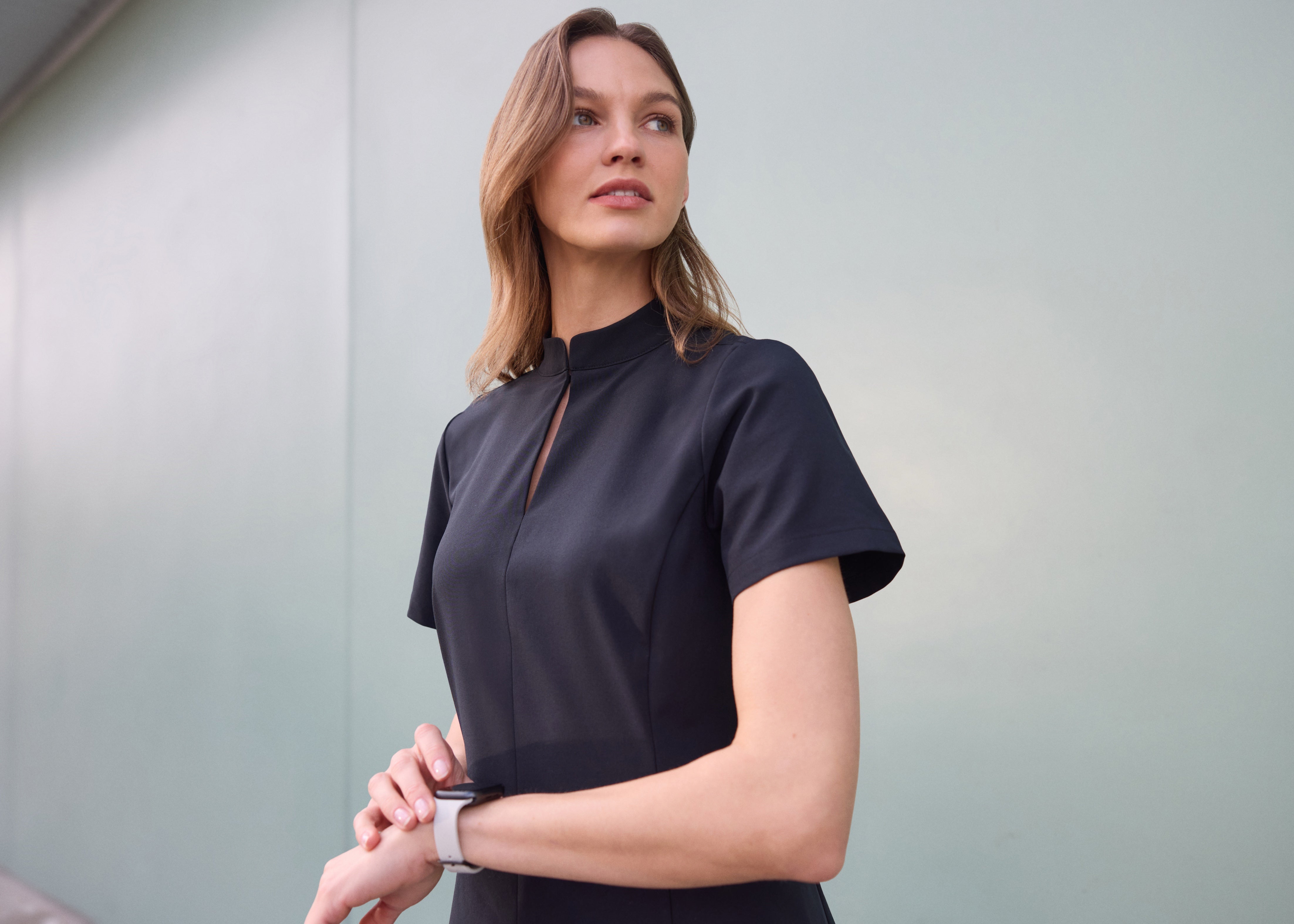 Woman in a black short-sleeve mock-neck scrub top with a keyhole opening, checking her smartwatch against a pale gray wall