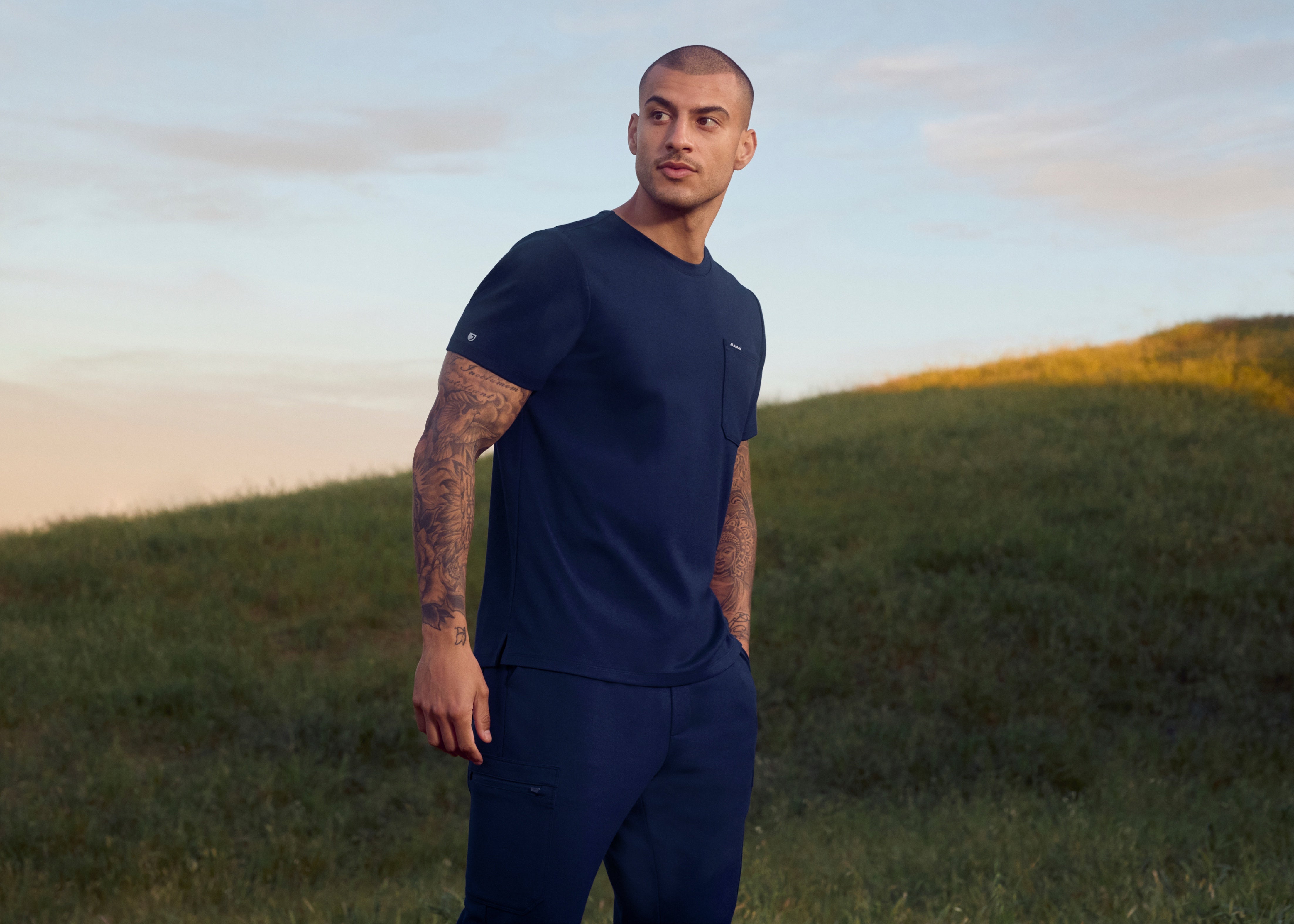 Man with tattooed arms in a navy crewneck scrub top with a chest pocket and matching cargo scrub pants, standing on a grassy hill at sunset