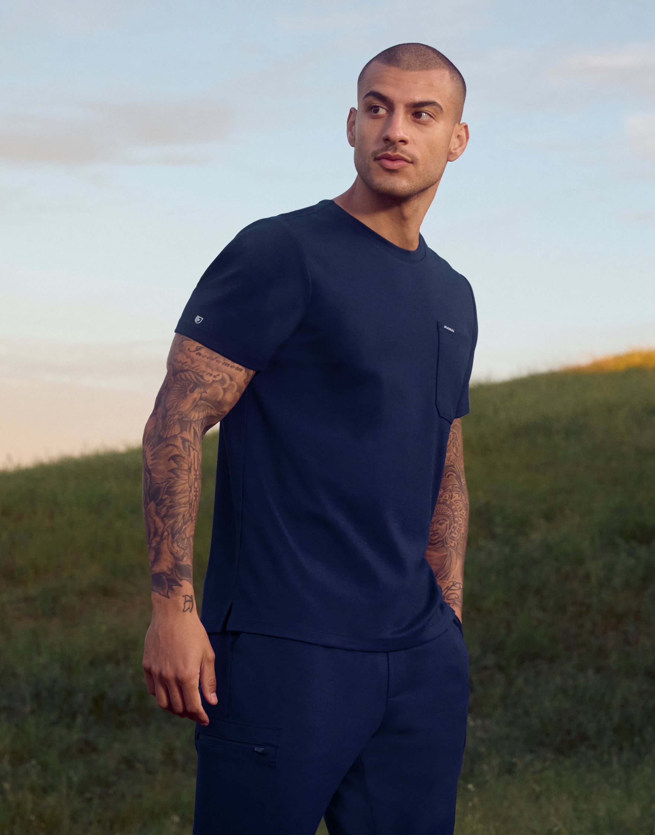 Man with tattooed arms in a navy crewneck scrub top with a chest pocket and matching cargo scrub pants on a grassy hill