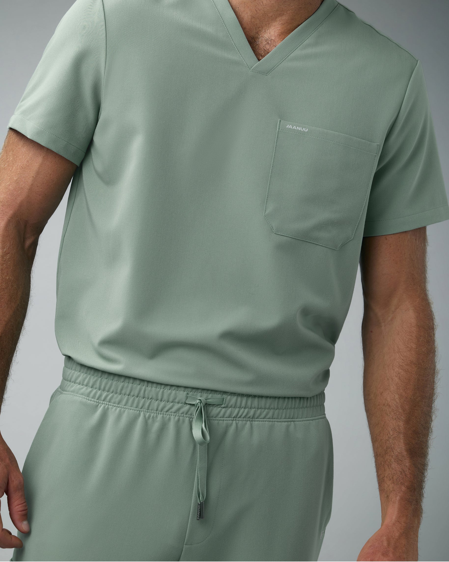Close-up of a sage green V-neck scrub top with JAANUU on the chest pocket over the drawstring waist of matching scrub pants