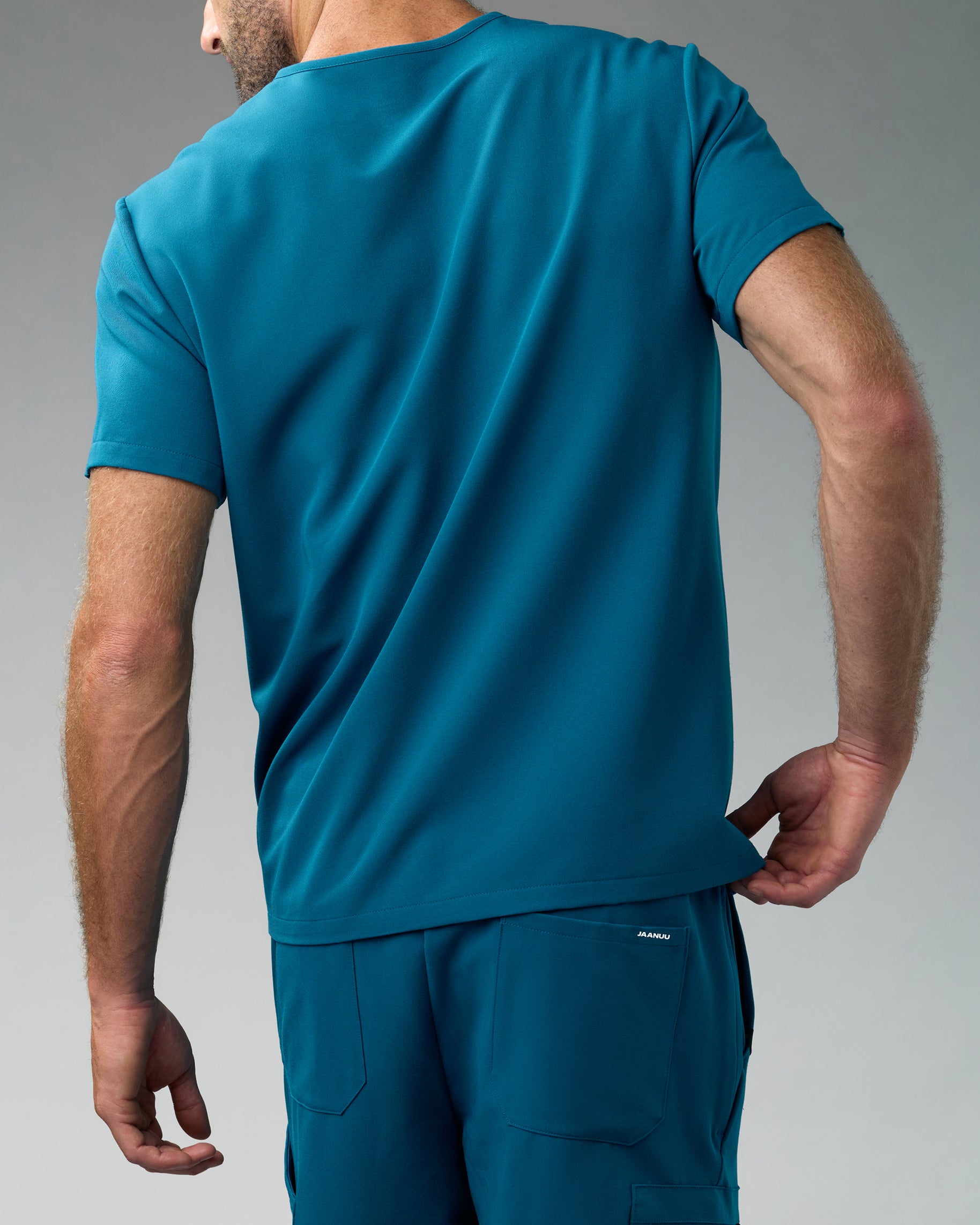 Back view of a man in a teal scrub top and matching scrub pants with a back pocket marked JAANUU