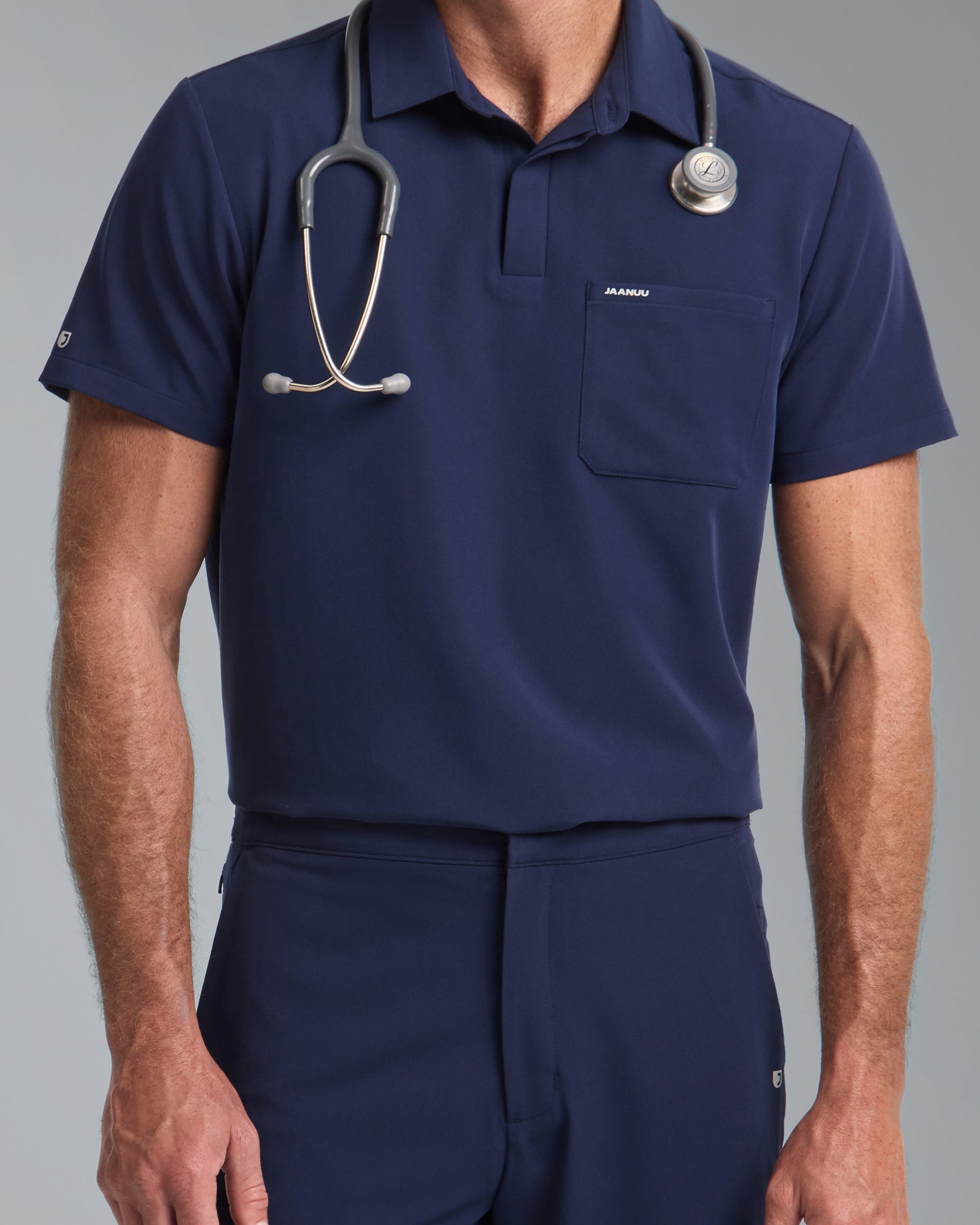 Man in a navy polo scrub top with a chest pocket marked JAANUU and a stethoscope around his neck, with matching scrub pants