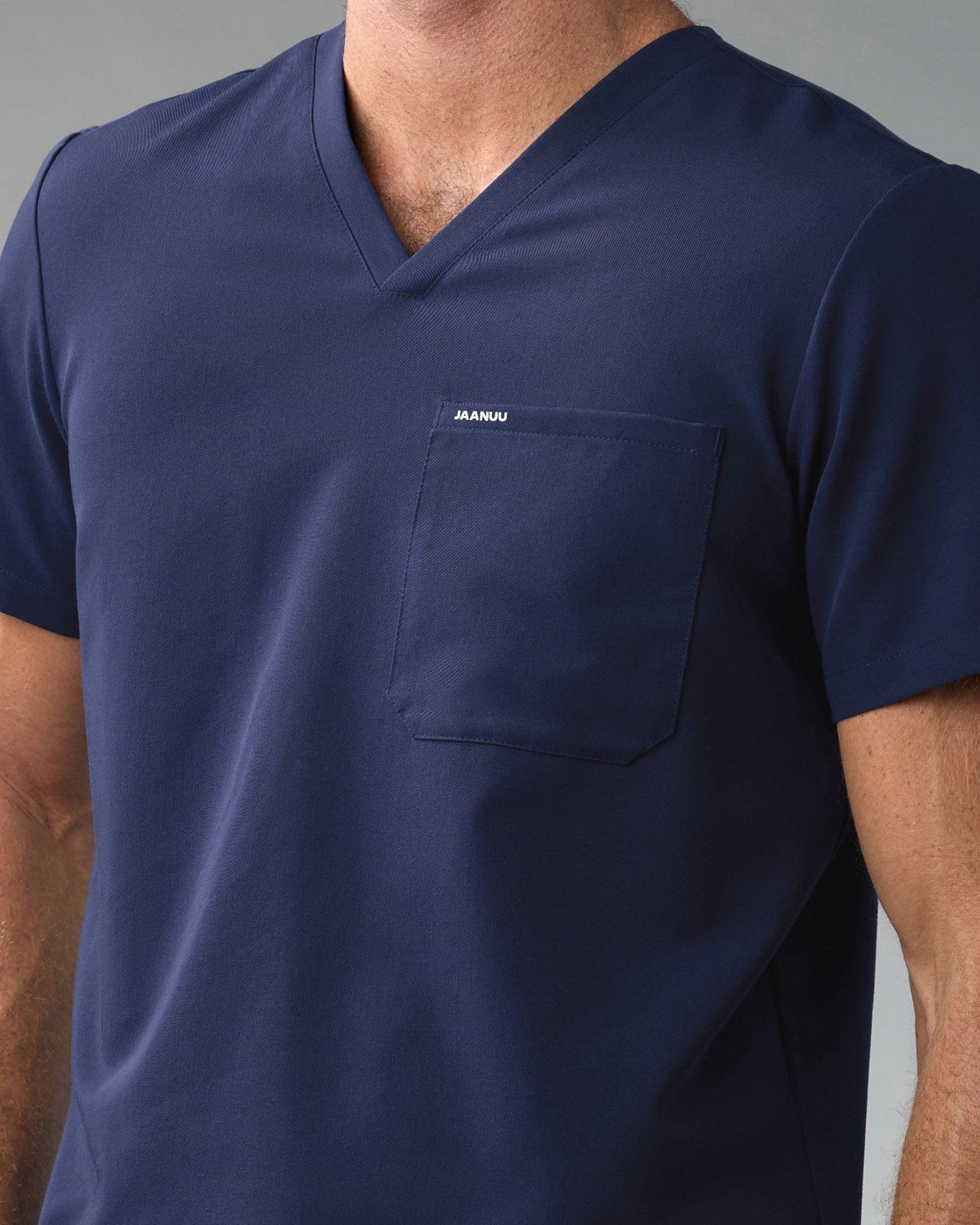 Close-up of the V-neckline and chest pocket marked JAANUU on a navy scrub top