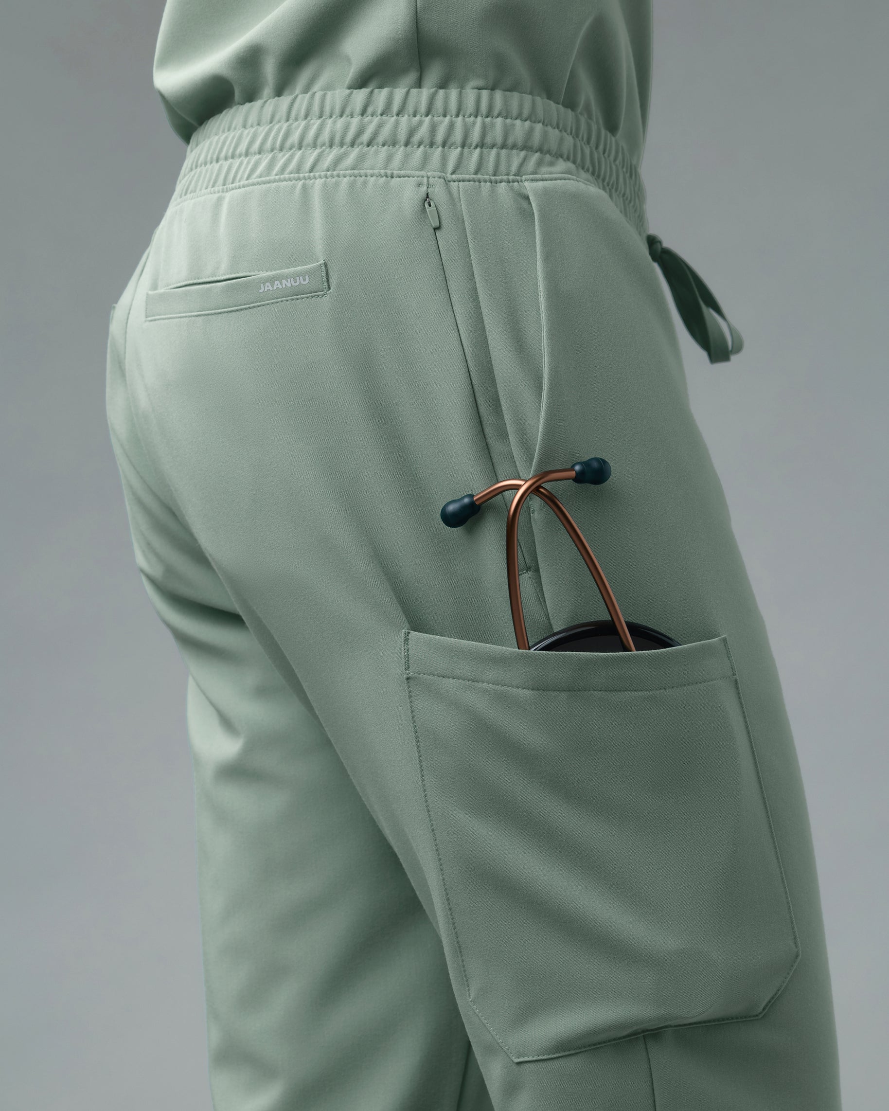 Close-up of a stethoscope in the cargo pocket of sage green scrub pants, with a zippered side pocket and a back welt pocket marked JAANUU