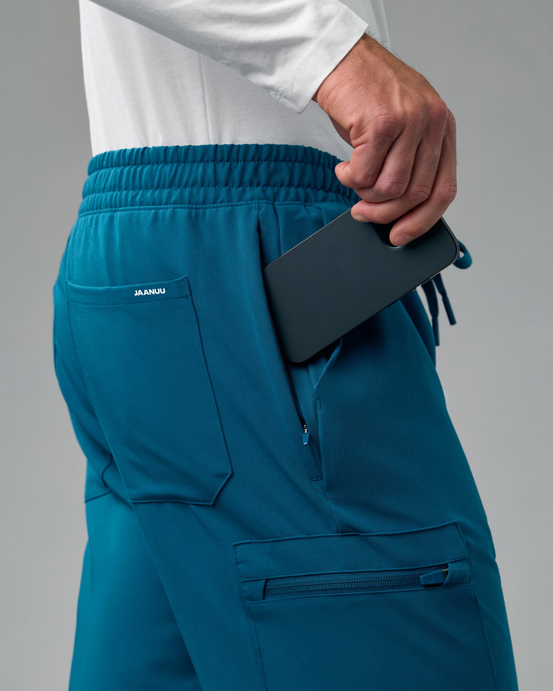 Close-up of a hand slipping a phone into the zippered side pocket of teal scrub joggers, with a back pocket marked JAANUU and a zippered cargo pocket