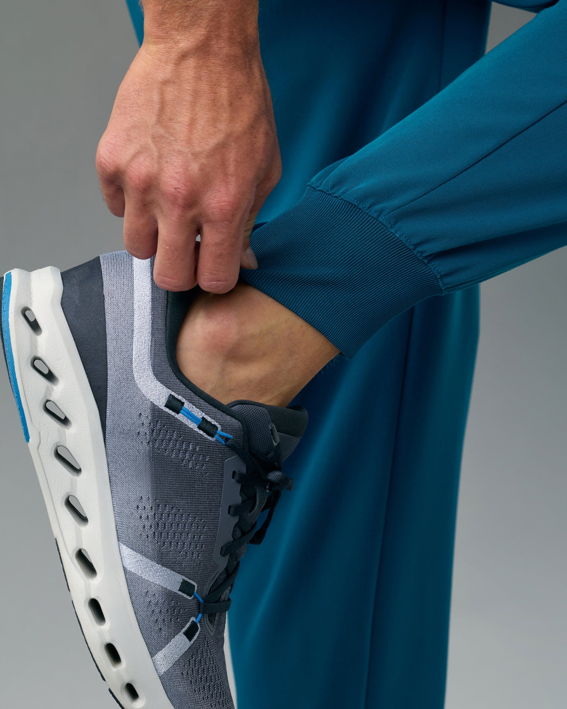 Close-up of a hand at the rib-knit ankle cuff of teal scrub joggers above a gray sneaker
