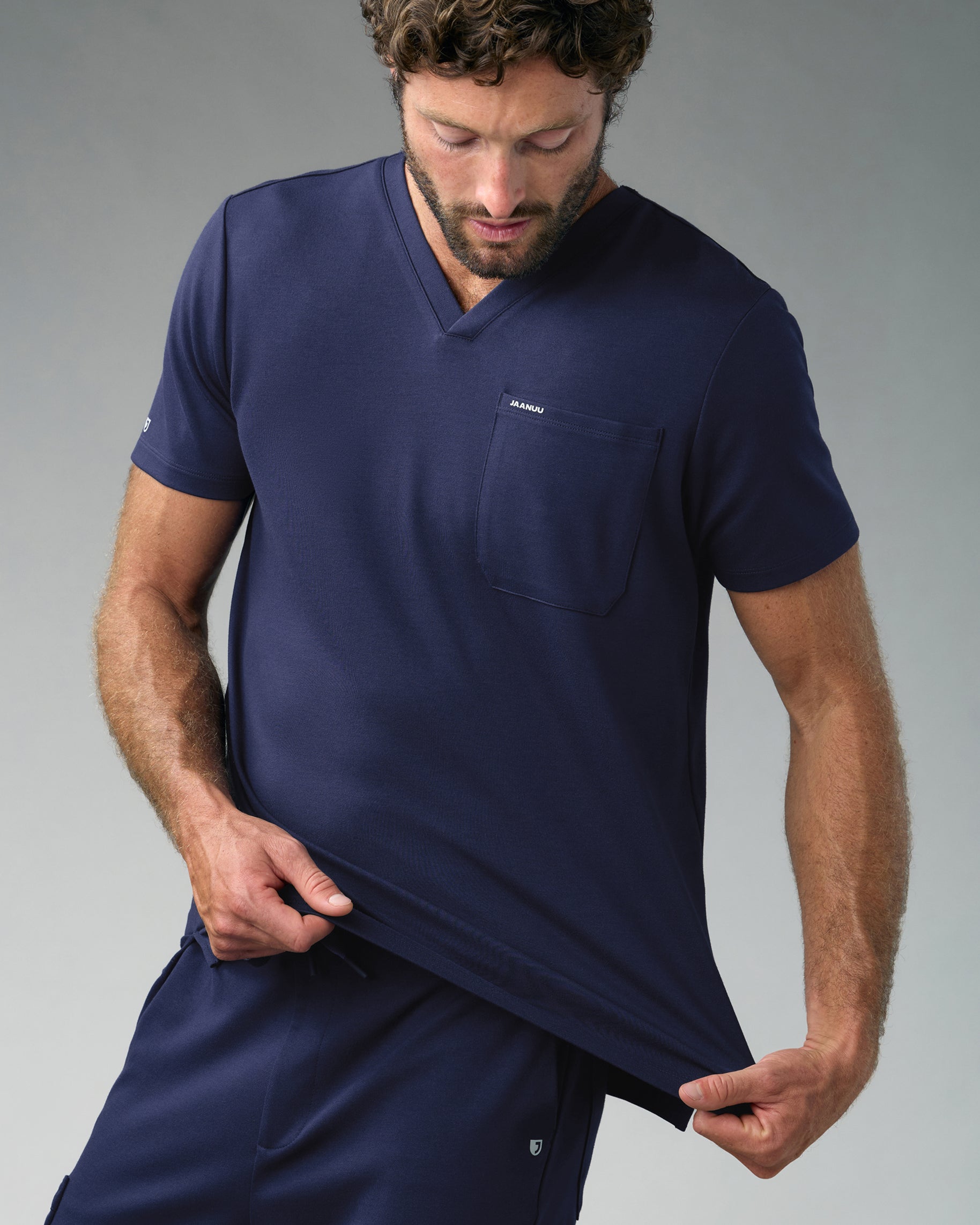 Man stretching the hem of a navy slim V-neck scrub top with JAANUU on the chest pocket