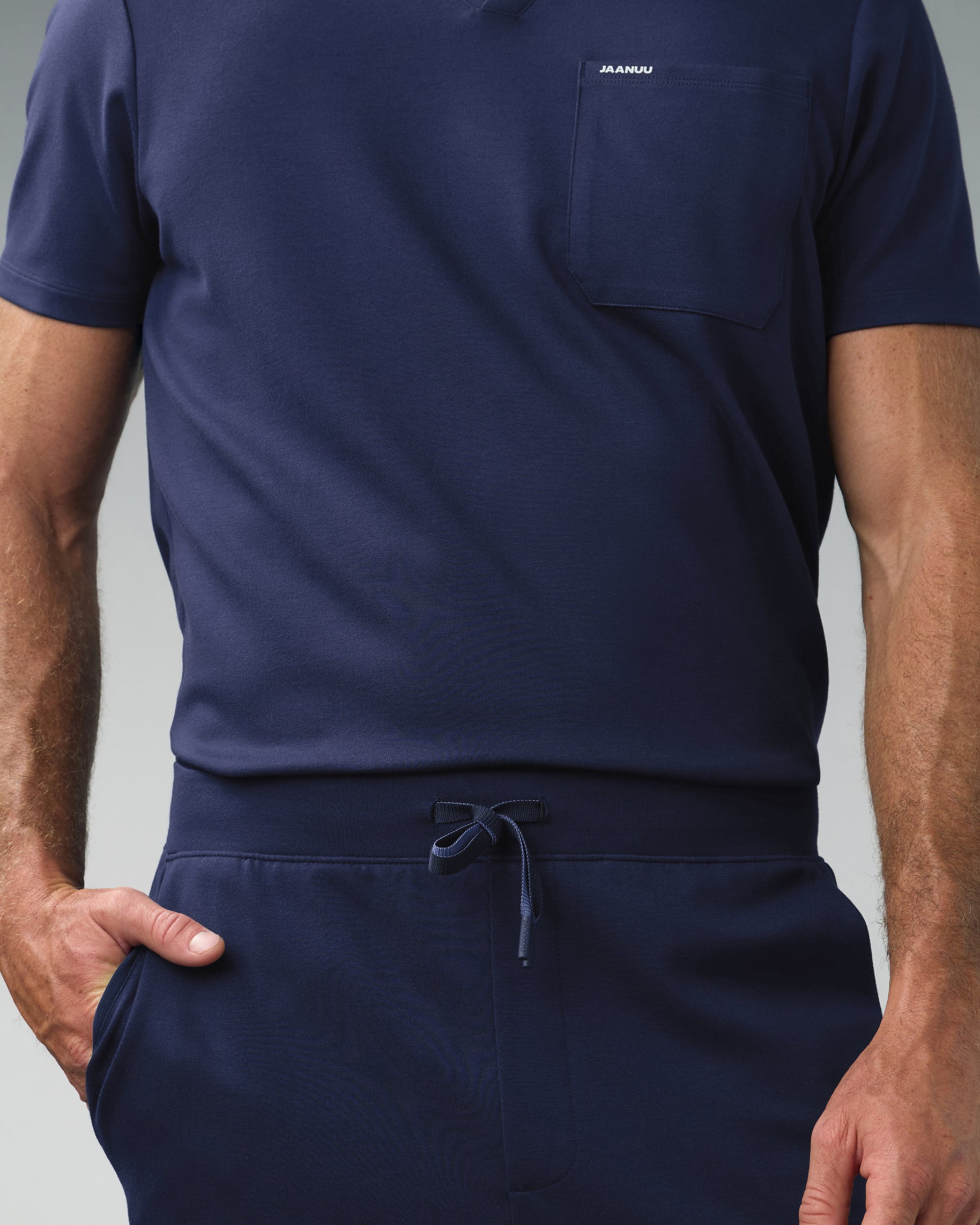 Close-up of a navy V-neck scrub top with JAANUU on the chest pocket over the drawstring waist of matching scrub pants
