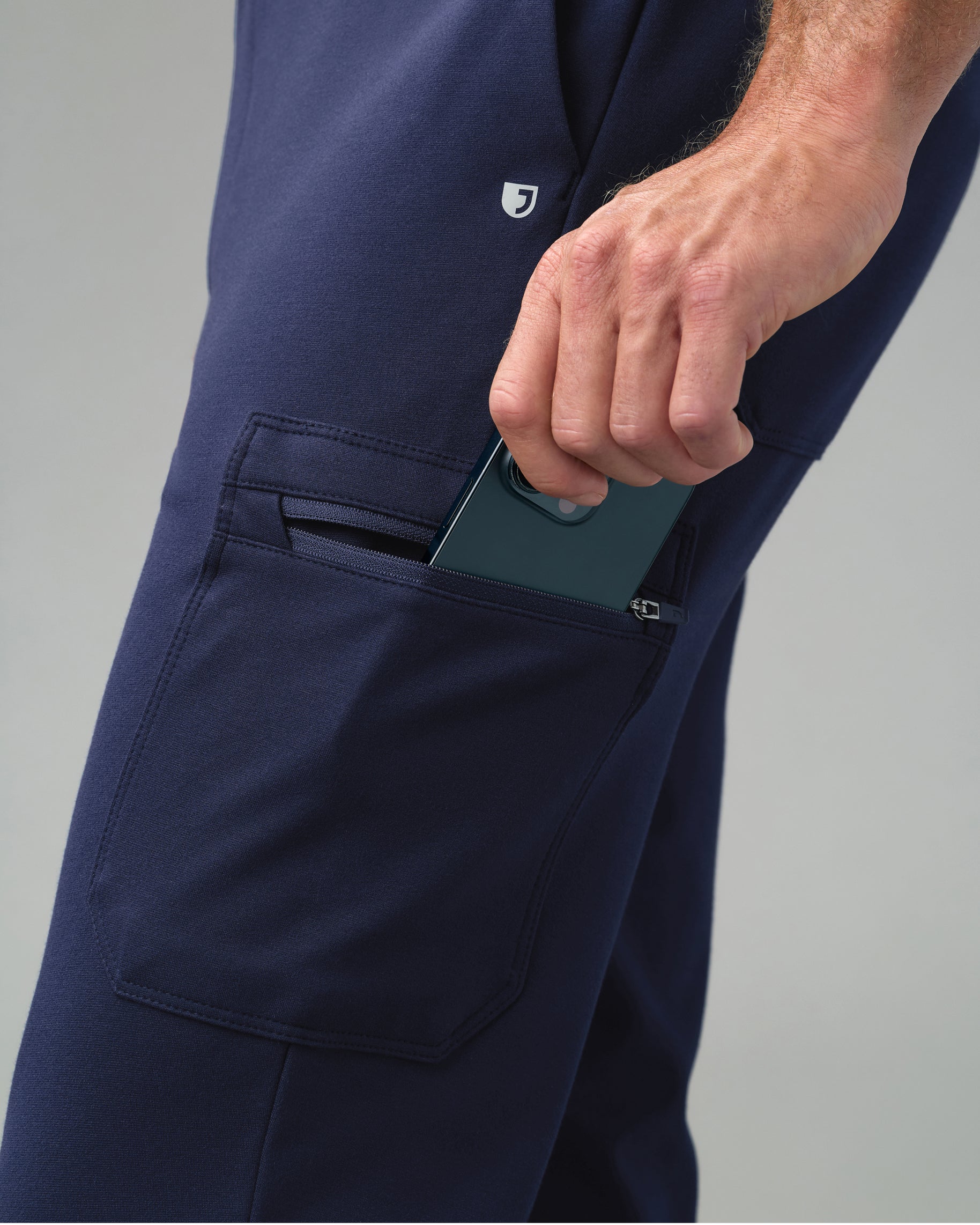 Close-up of a hand slipping a phone into the zippered cargo pocket of navy slim scrub joggers