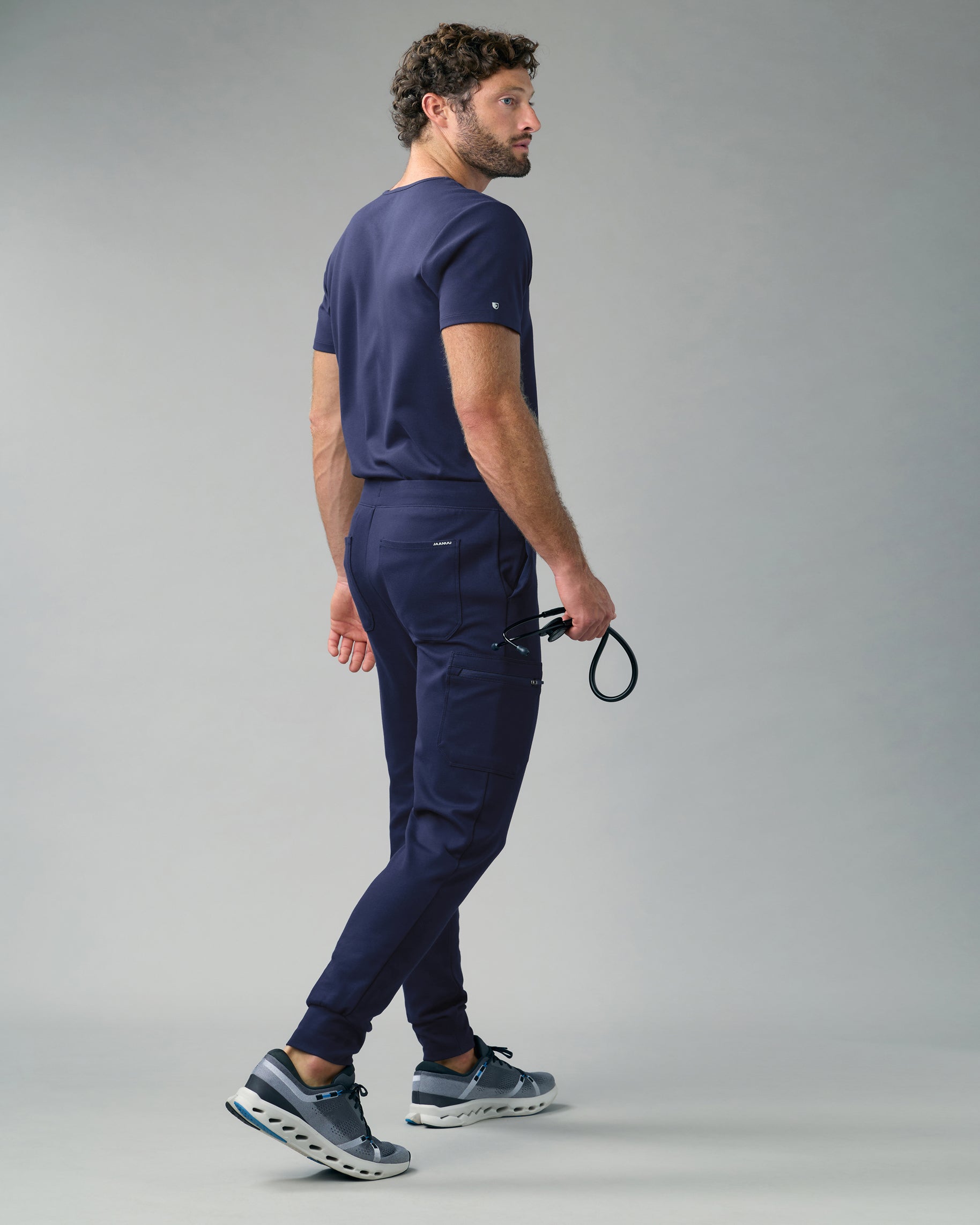 Back view of a man in a navy scrub top and slim cargo scrub joggers, holding a stethoscope