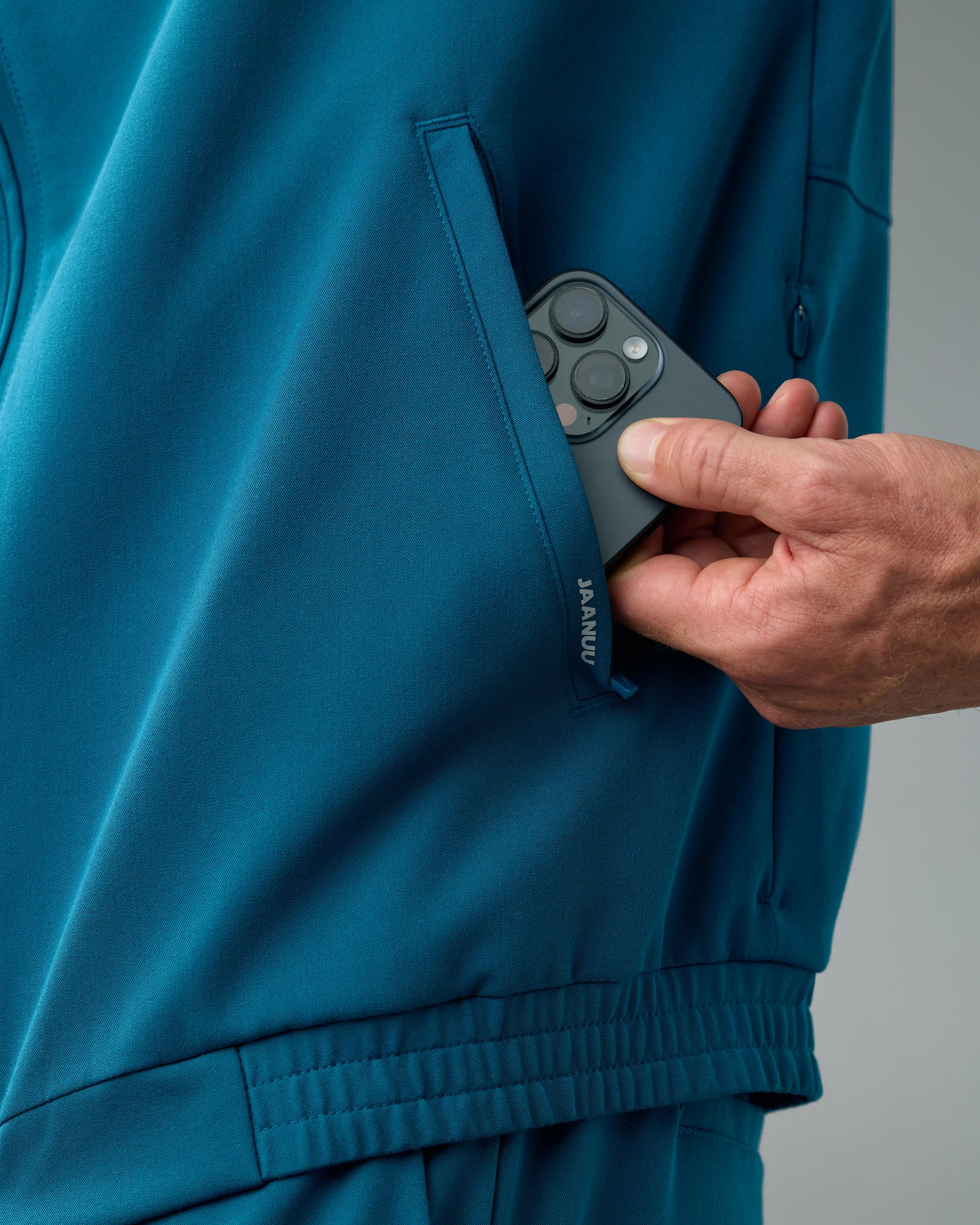 Close-up of a hand slipping a phone into the side pocket marked JAANUU on a teal scrub jacket