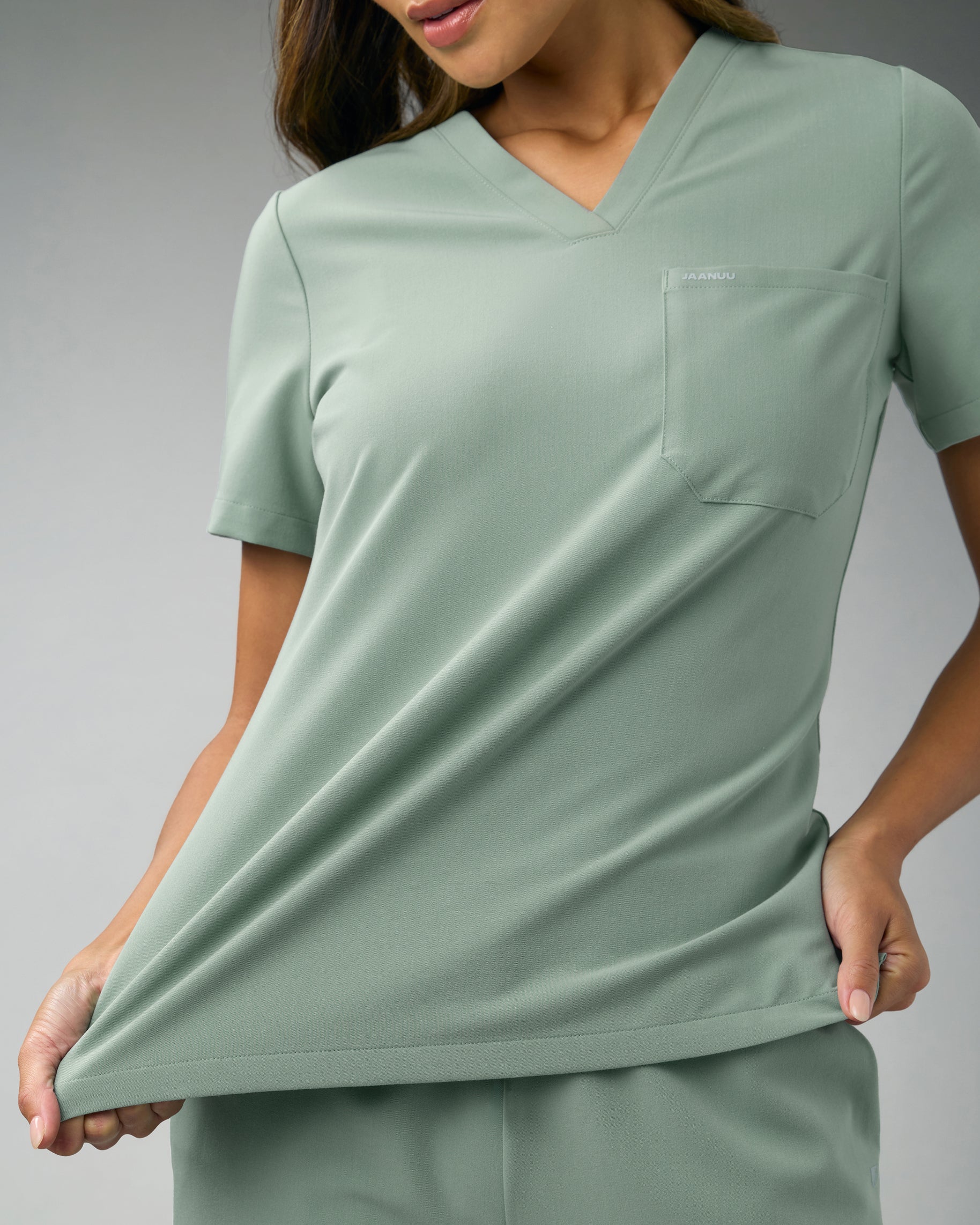 Woman stretching the hem of a sage green V-neck scrub top with a chest pocket to show its stretch