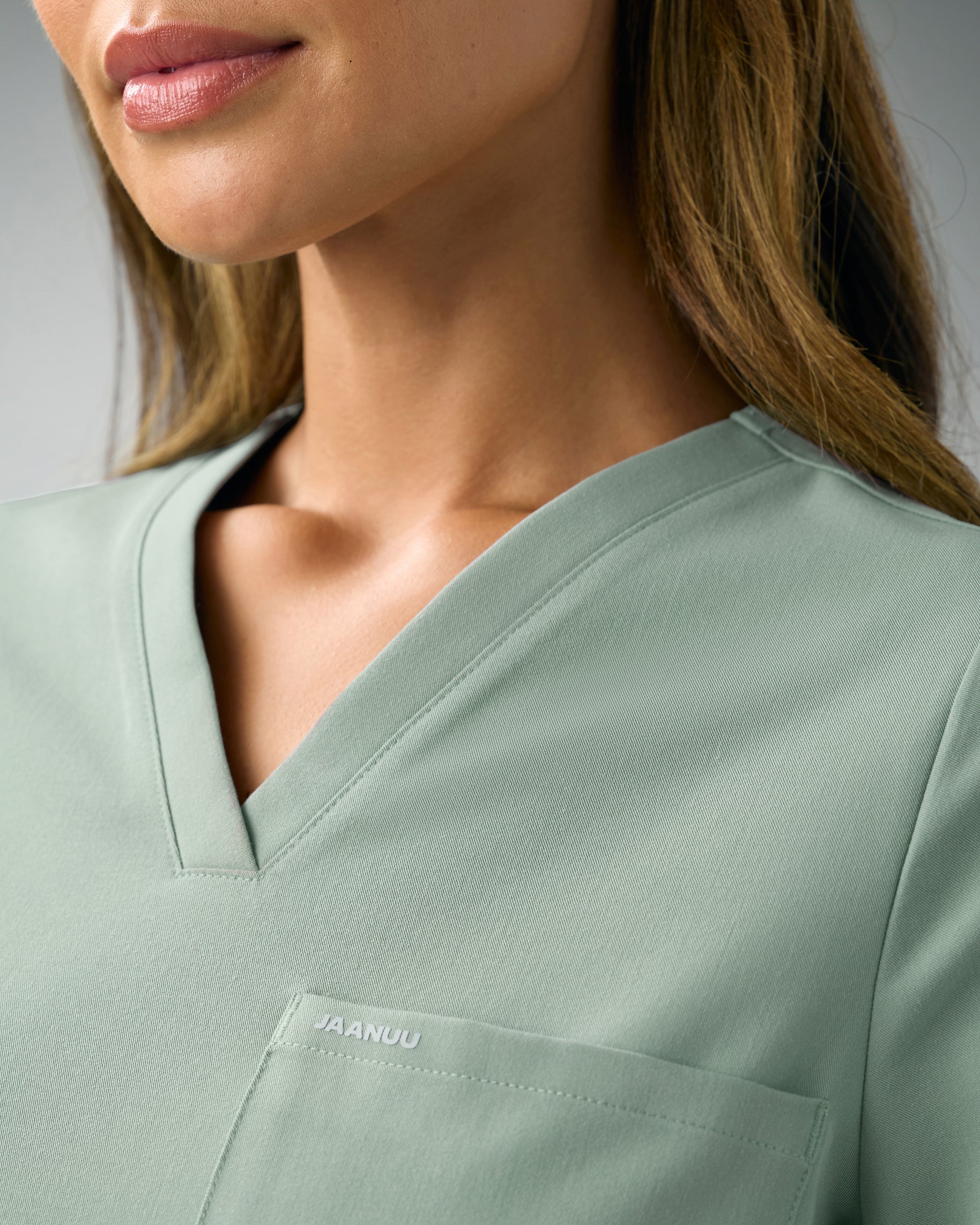Close-up of the V-neckline of a sage green scrub top with JAANUU on the chest pocket