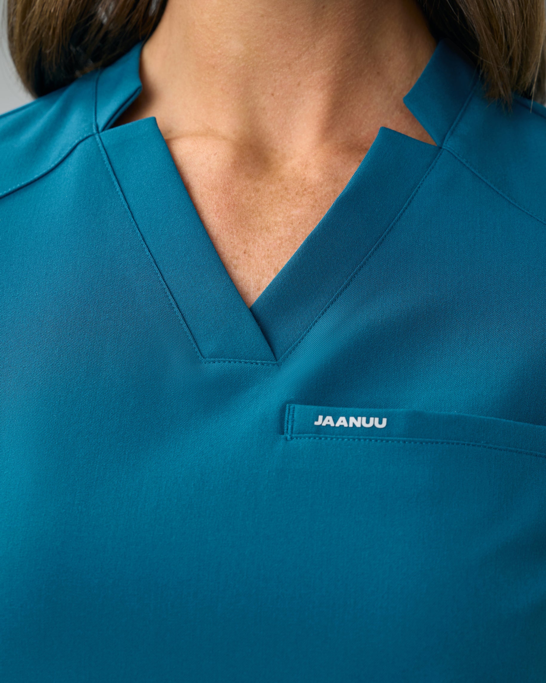 Close-up of the notched V-neckline of a teal scrub top with JAANUU on the chest welt pocket