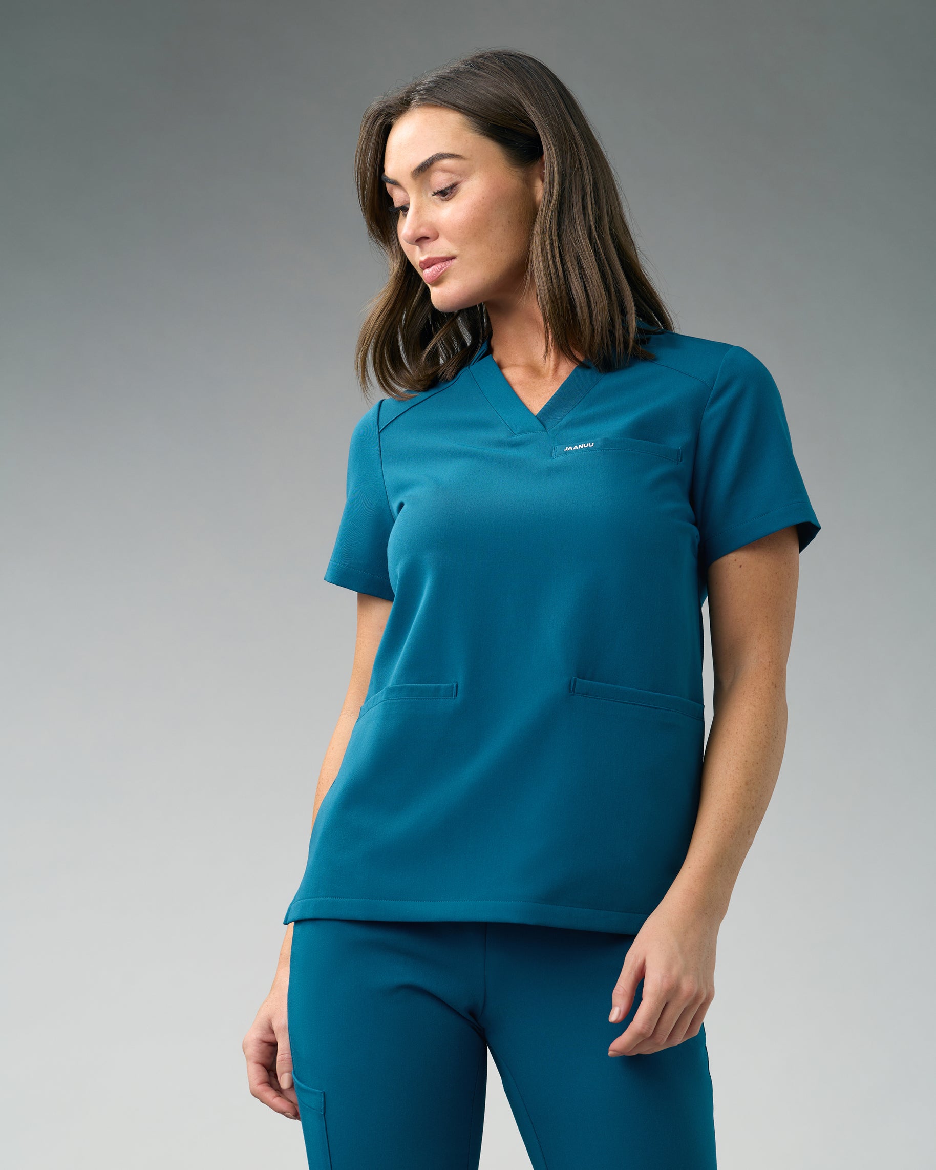 Woman in a teal notched V-neck scrub top with a chest welt pocket, two front welt pockets and matching scrub pants