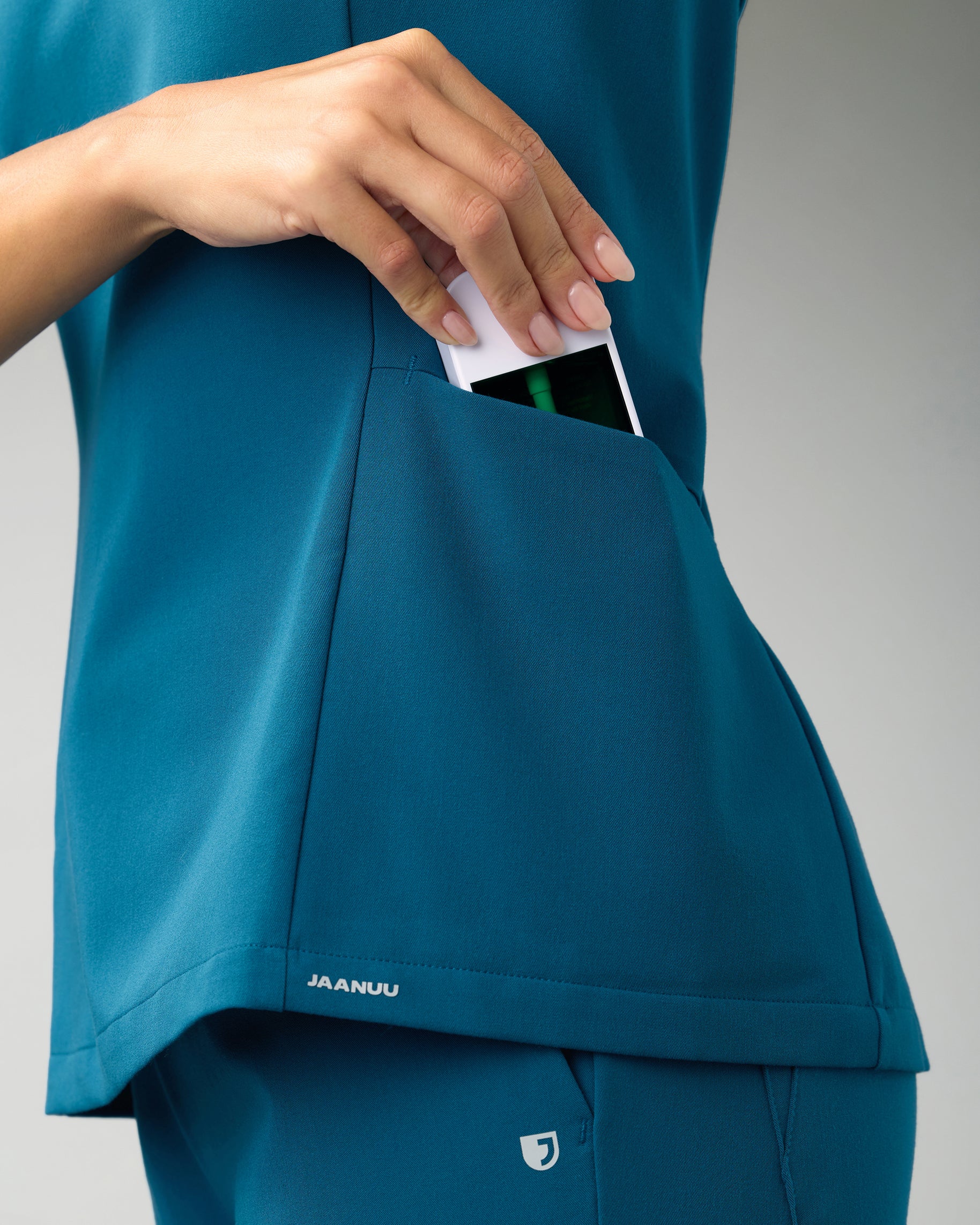 Close-up of a hand slipping a device into the side pocket of a teal scrub top with a JAANUU logo at the hem