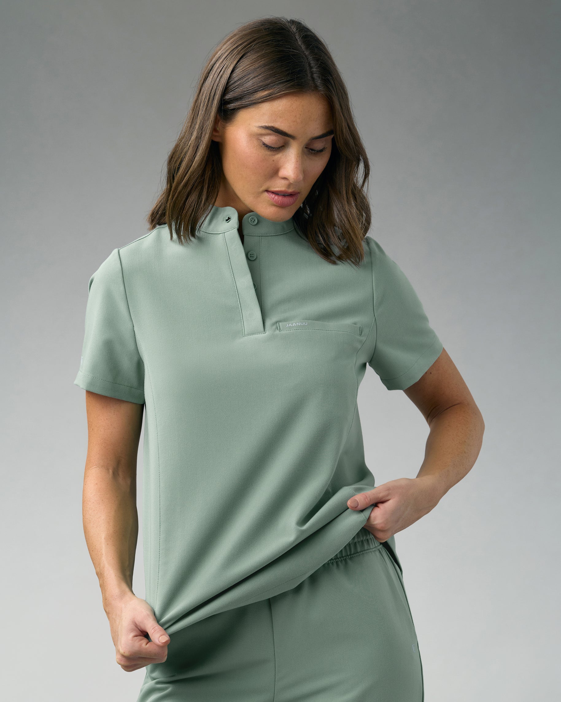 Woman in a sage green stand-collar scrub top pulling at the hem to show its stretch