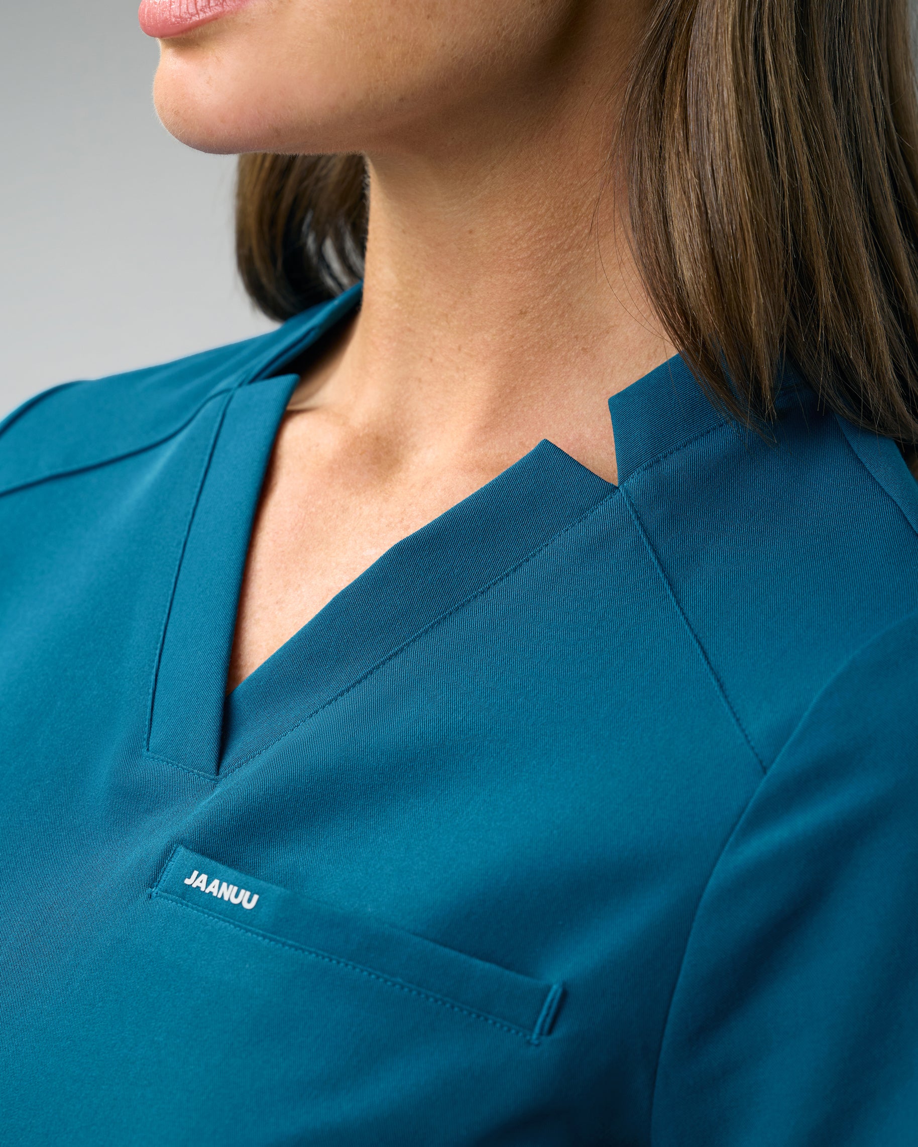 Close-up of the notched V-neckline of a teal slim scrub top with JAANUU on the chest welt pocket