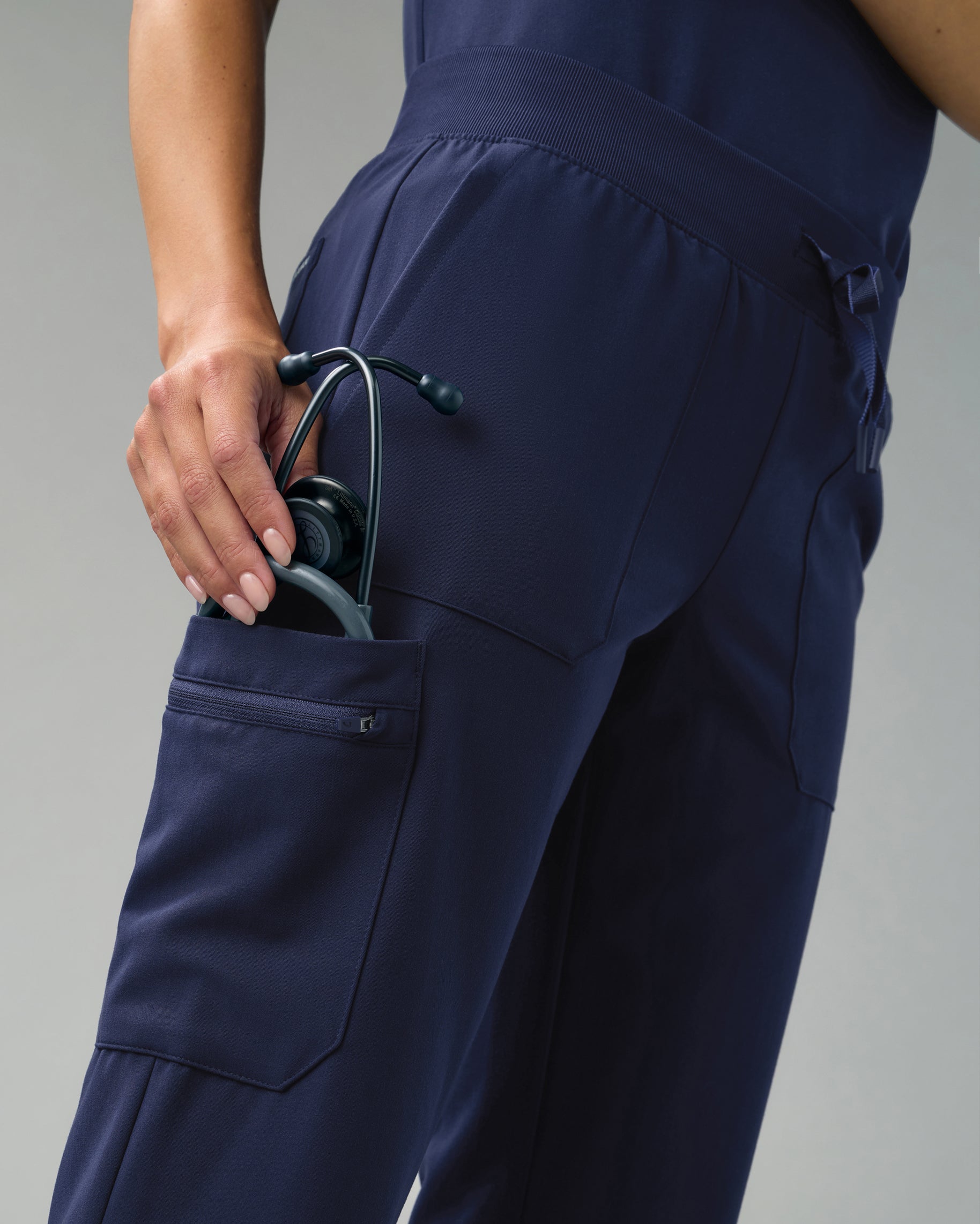Close-up of a hand tucking a stethoscope into the zippered cargo pocket of navy scrub pants