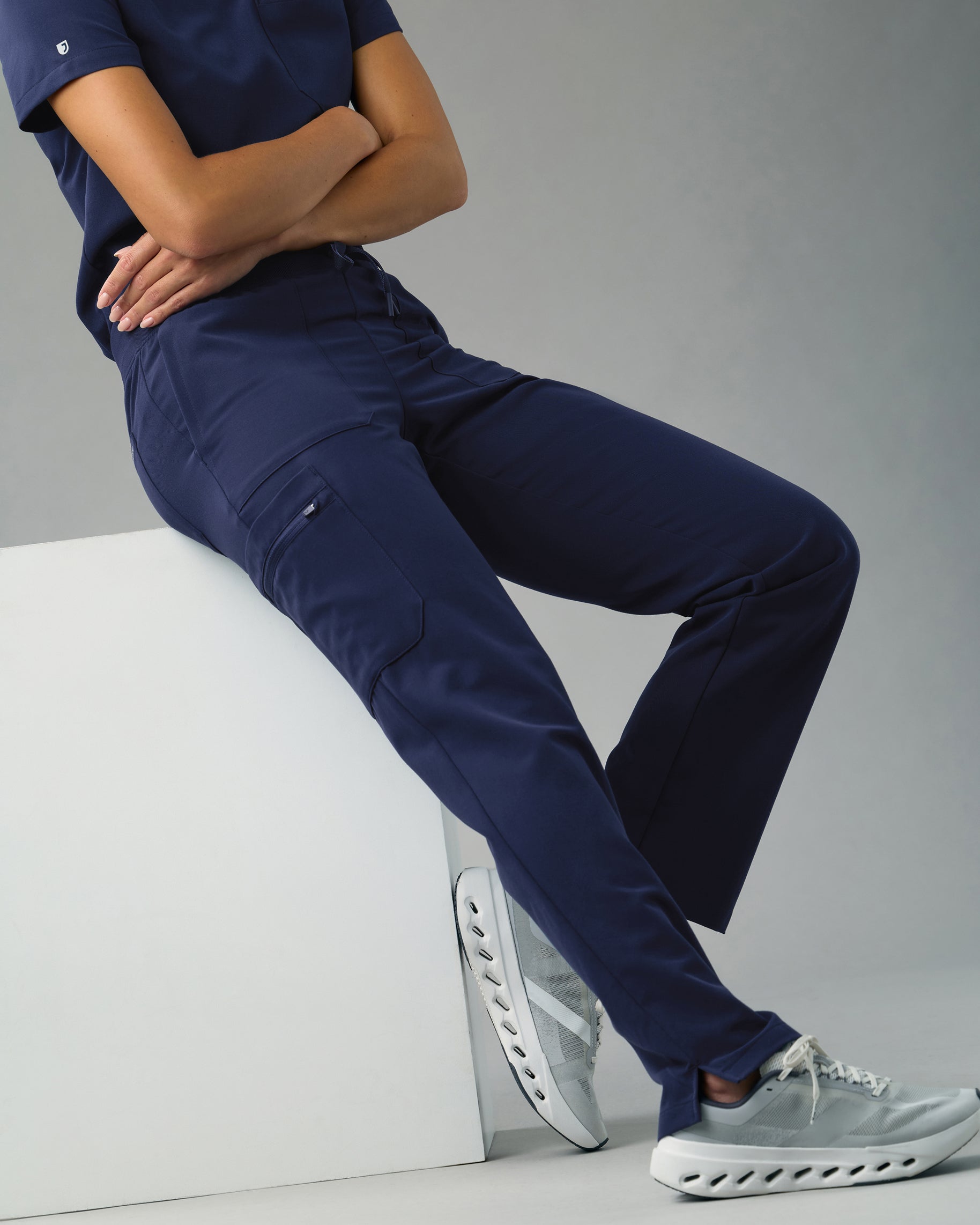 Woman sitting on a white block in navy cargo scrub pants with a zippered cargo pocket, arms crossed